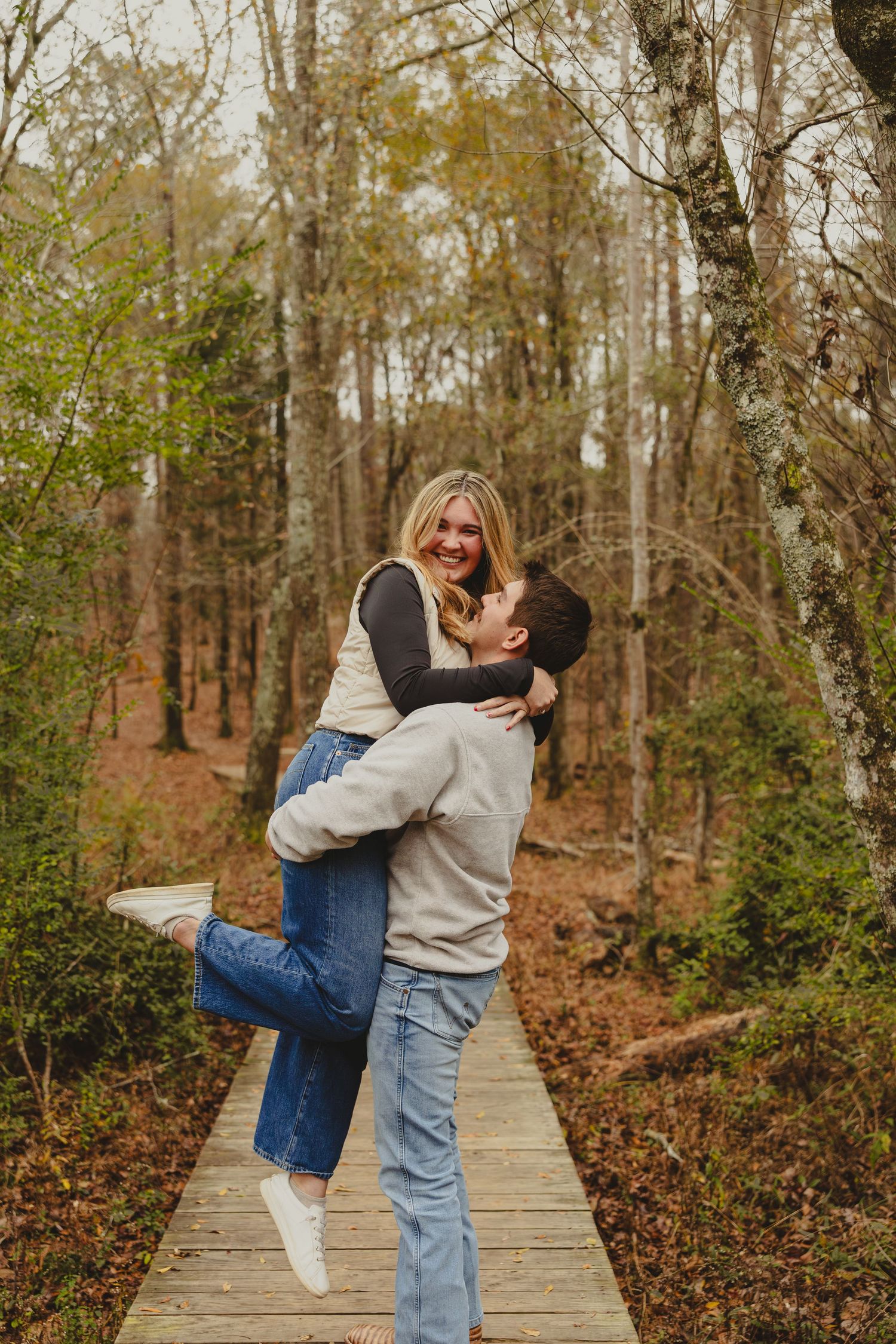 A couple playfully embraces and spins around on a wooden boardwalk path through an autumn forest.