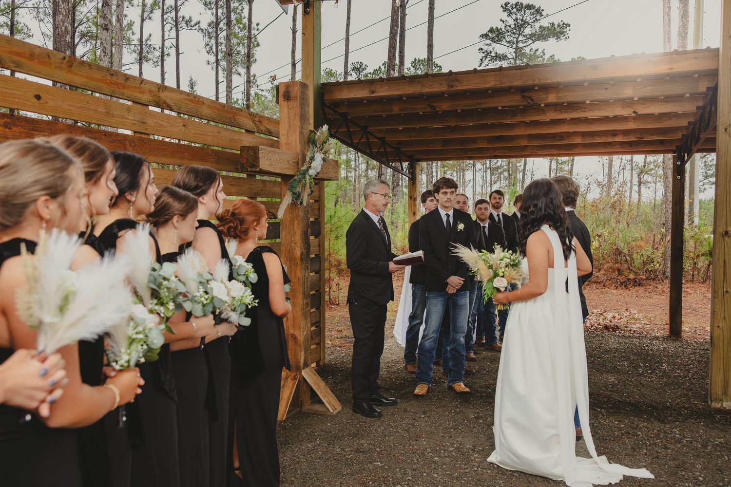 Wedding ceremony taking place under a rustic wooden pavilion with bridesmaids in black dresses holding white bouquets.