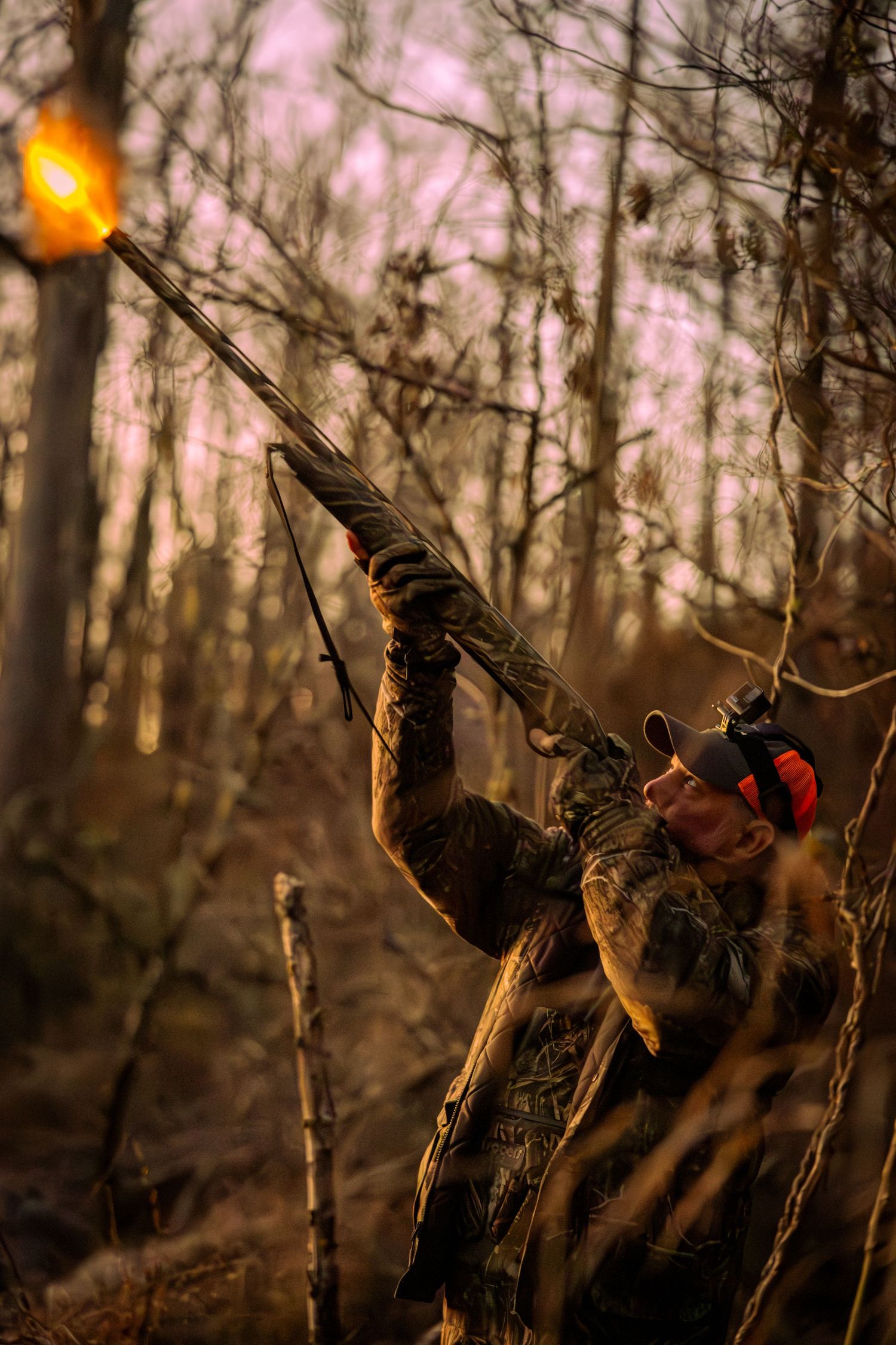 Hunter in camouflage gear aiming upward with a weapon in a misty forest at sunset.