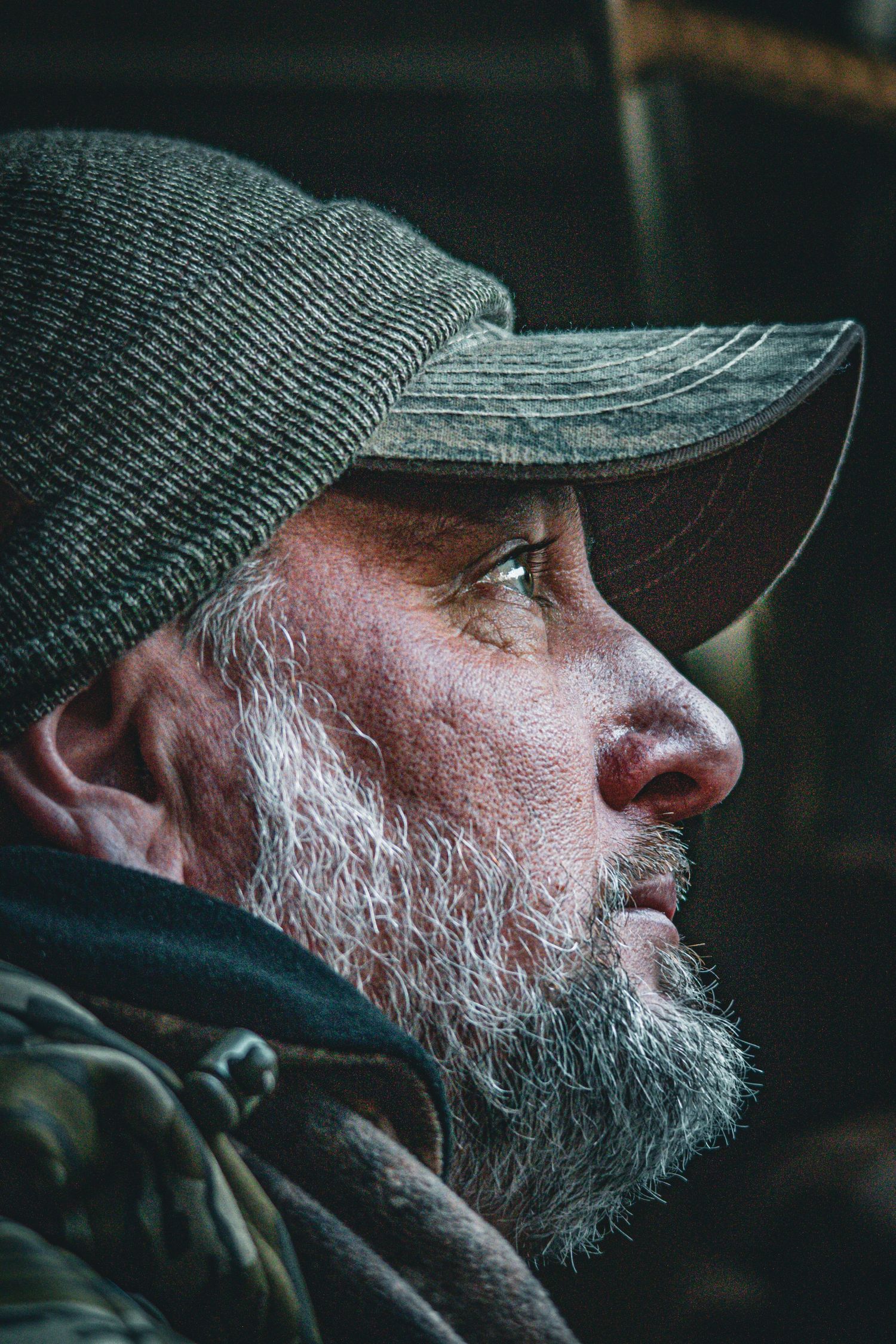 Side profile of person wearing dark knit cap with gray beard in outdoor setting.