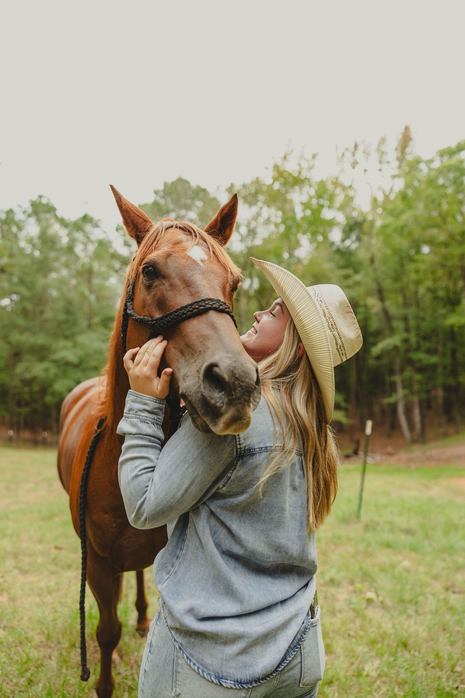Person in denim and hat sharing affectionate moment with brown horse in grassy field.