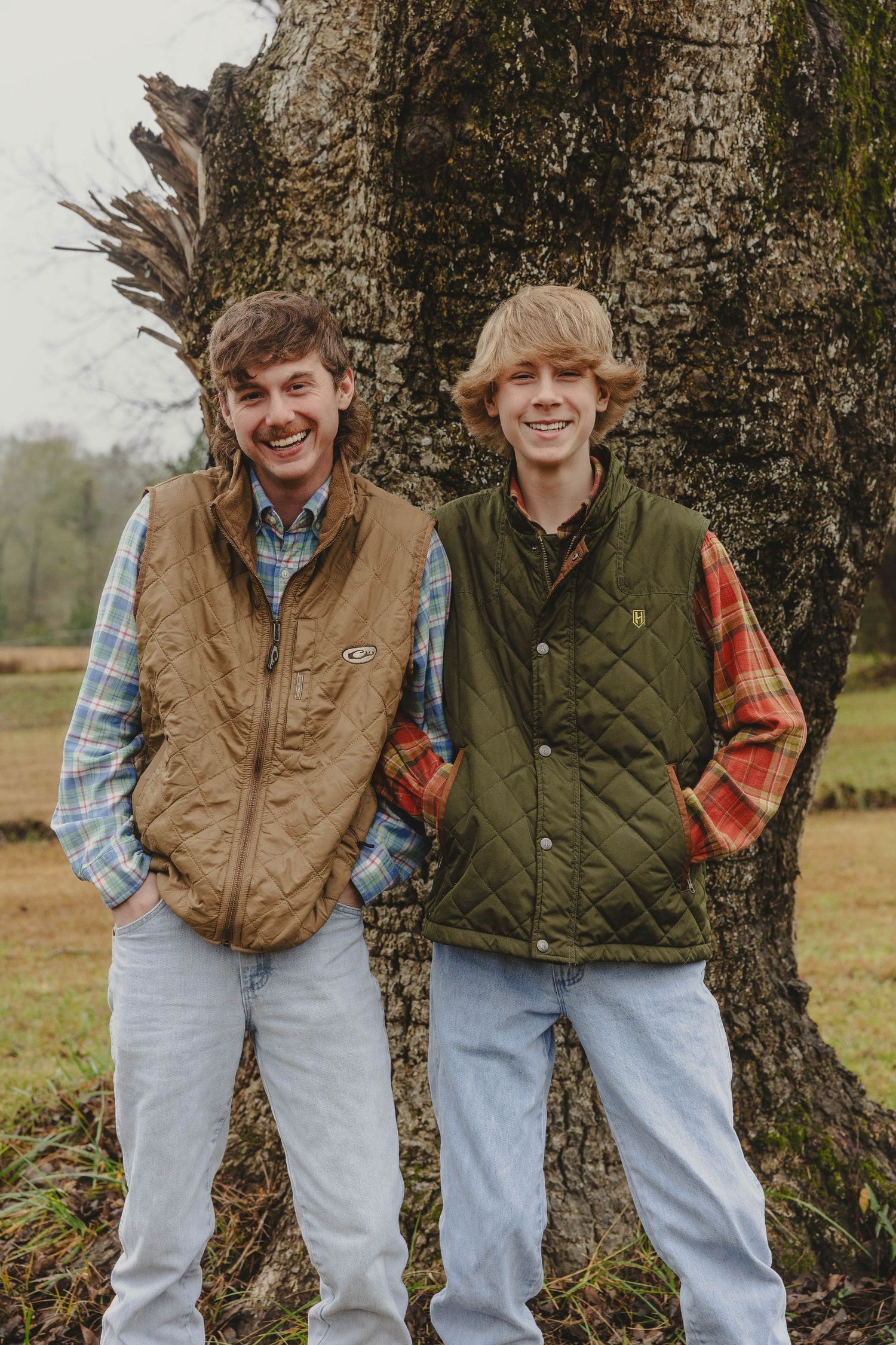 Two people in casual fall attire stand against a tree trunk wearing vests and plaid shirts.