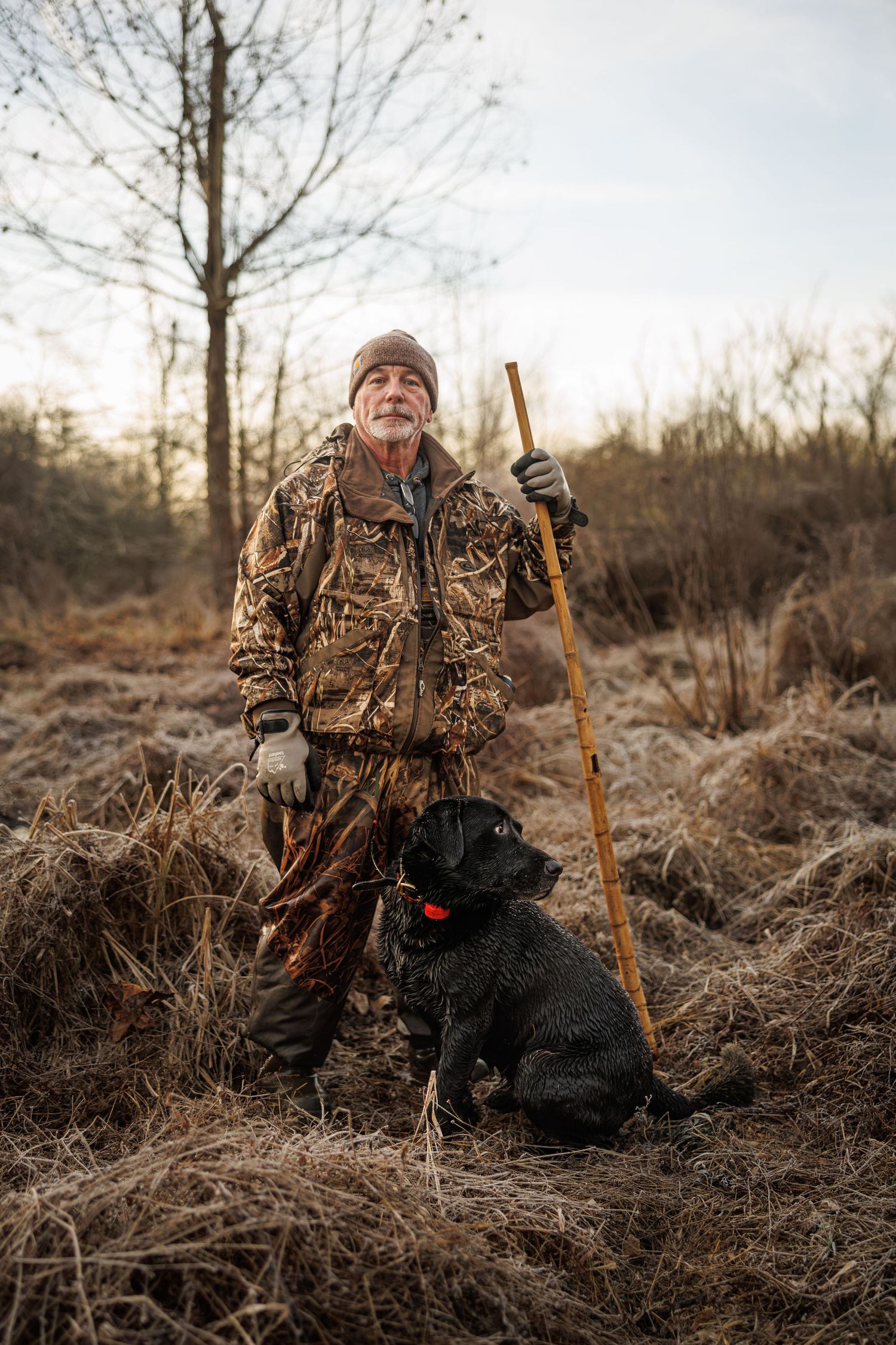 Hunter in camo clothing stands with walking stick and black hunting dog in autumn field.