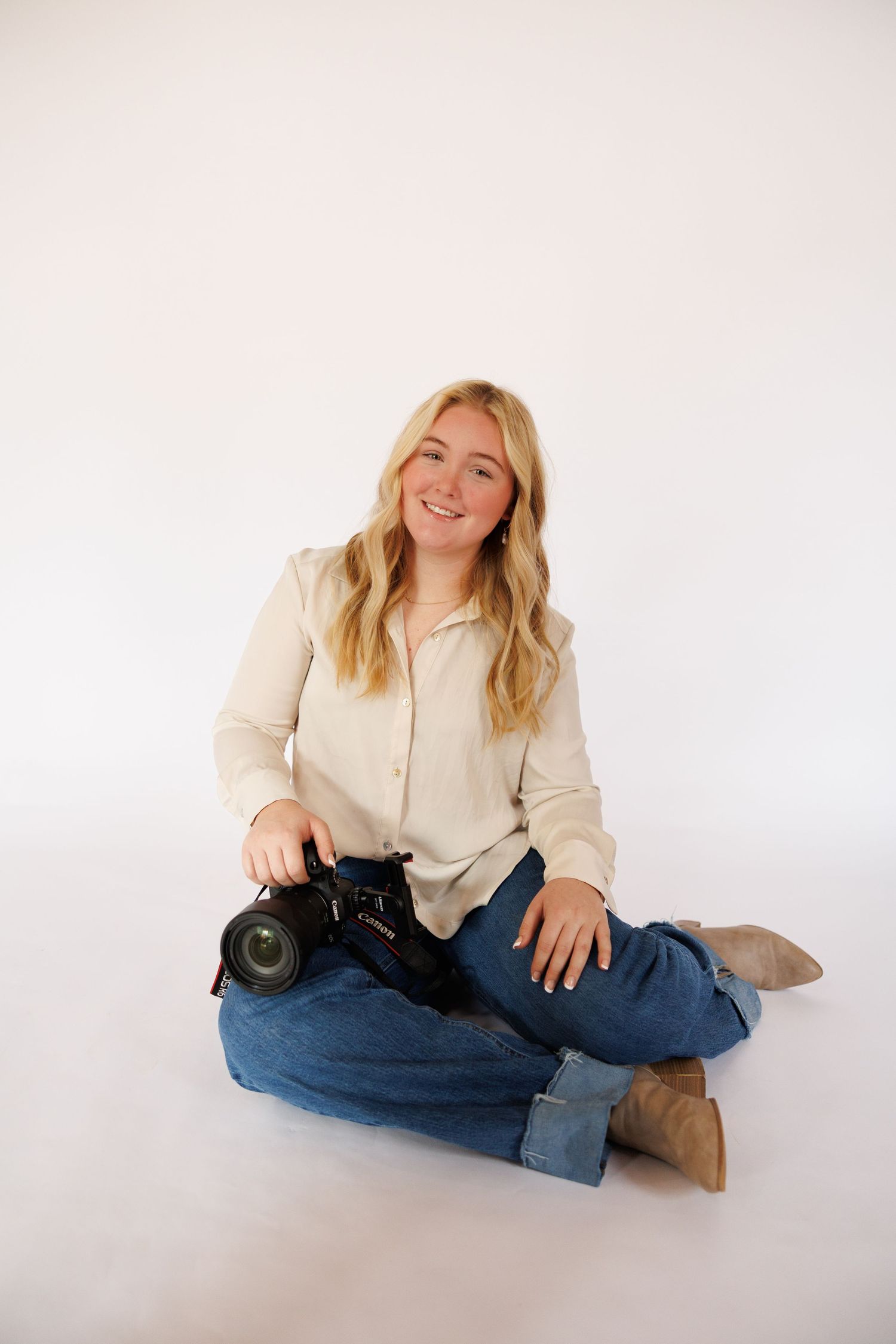 Photographer in cream sweater and jeans sits casually with camera on white backdrop in lifestyle photoshoot series.