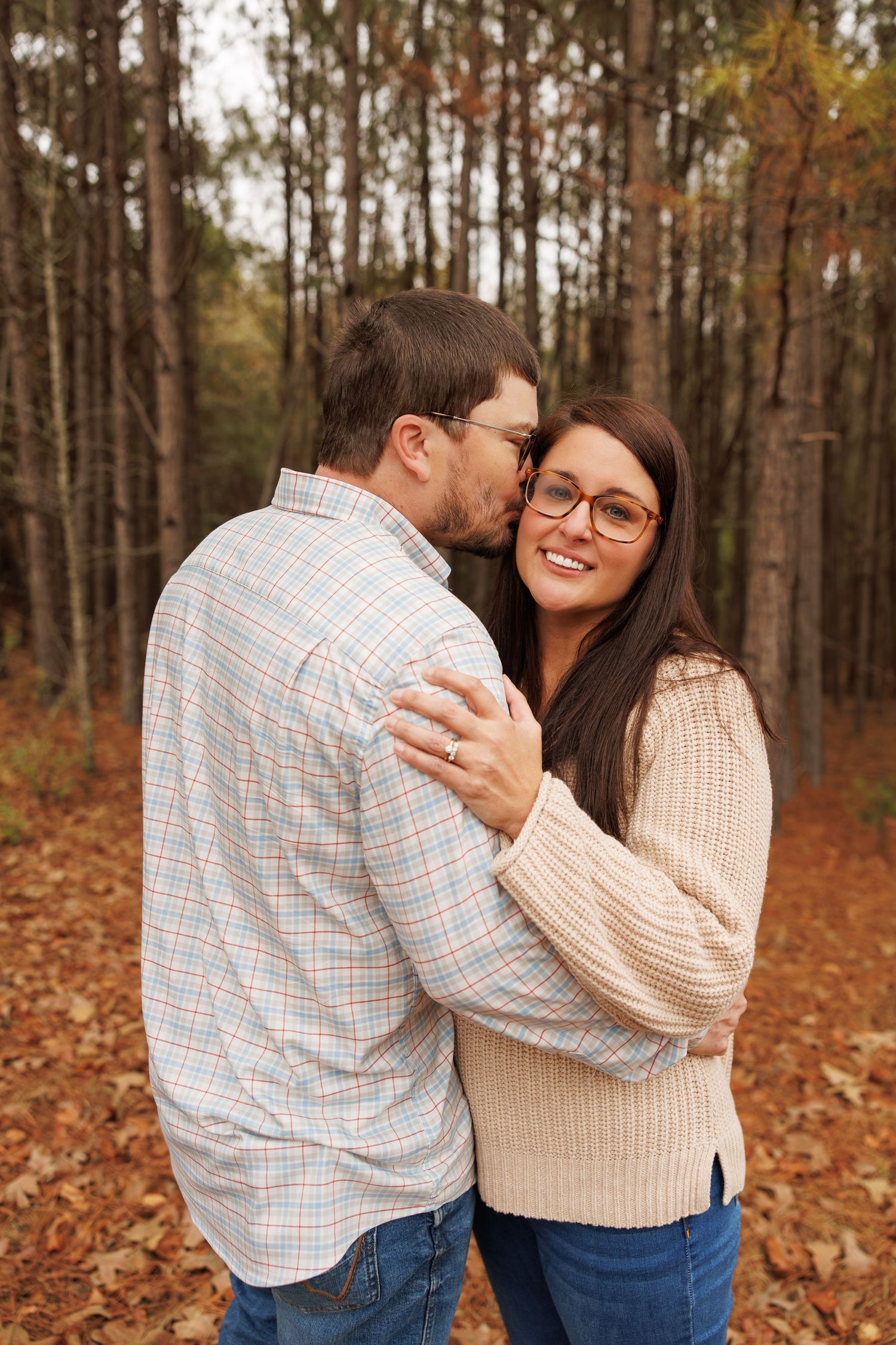 A couple embraces in a romantic autumn forest setting with fallen leaves and tall pine trees in the background.