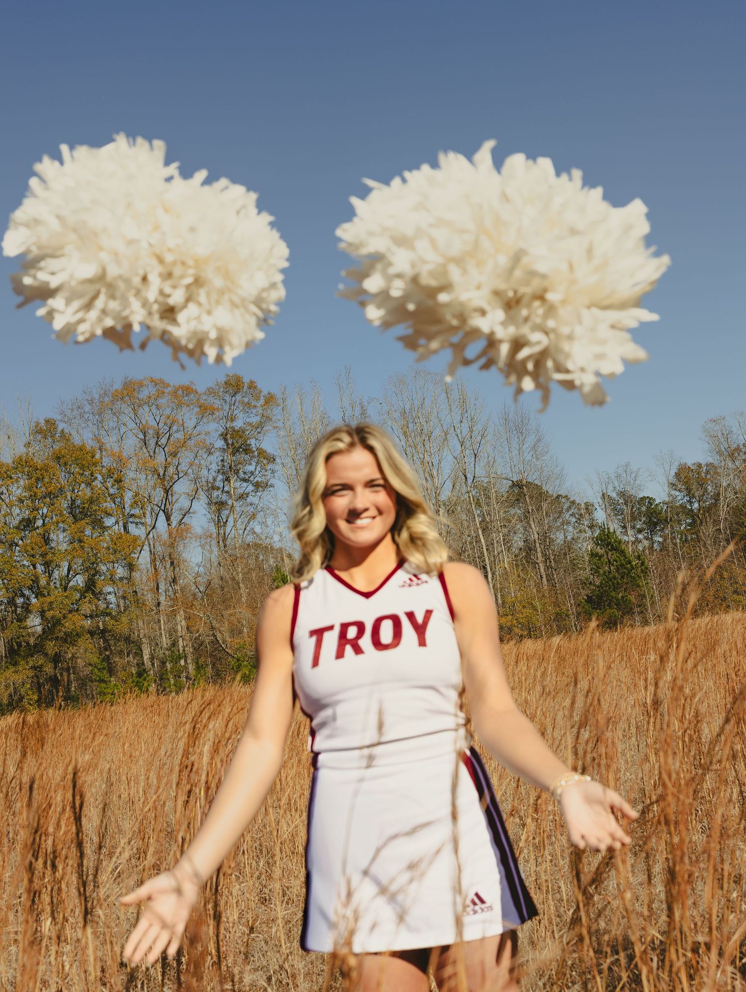A person in a white Troy sports jersey poses playfully in a grassy field on a sunny autumn day.