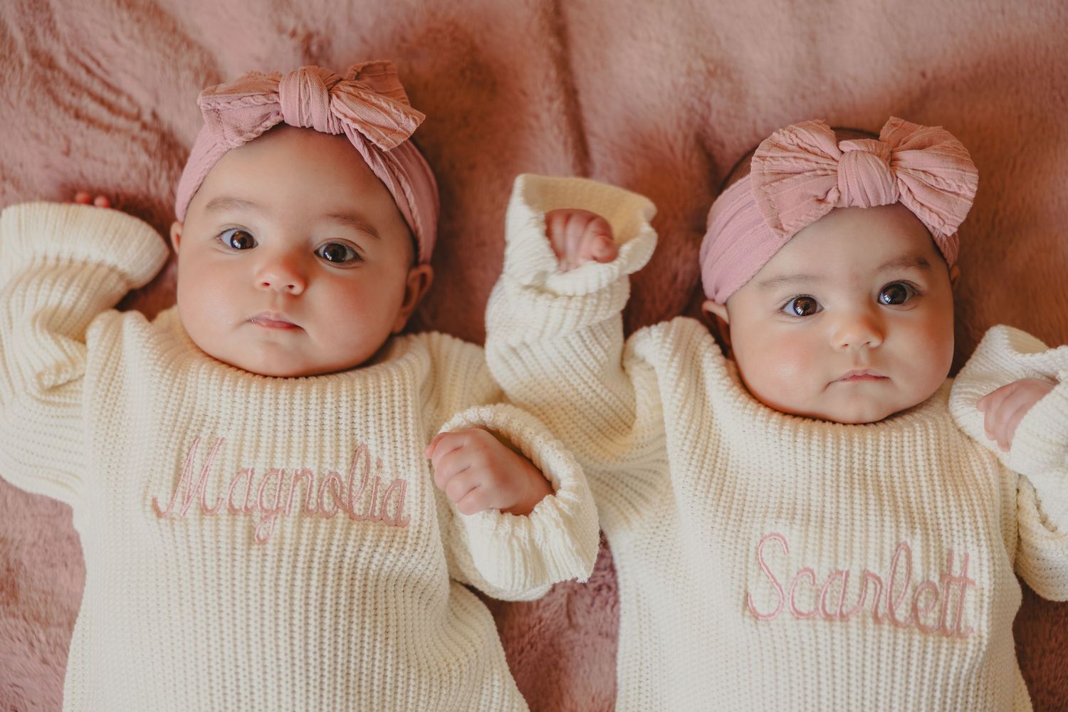 A baby wearing a cream knit sweater and pink bow lies on a pink blanket while looking at the camera.