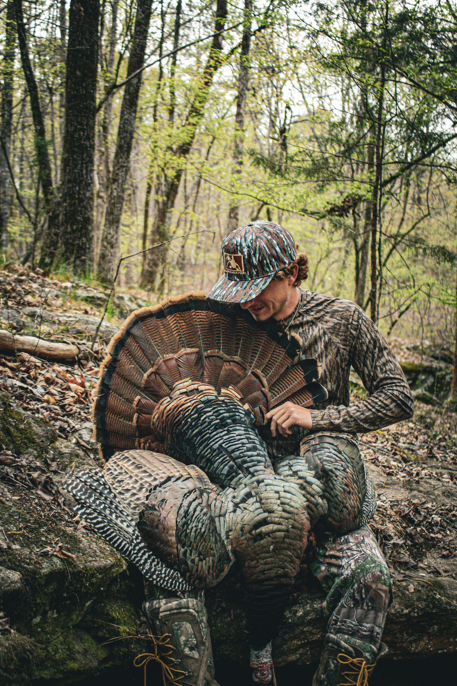 Hunter in camouflage examining harvested wild turkey in forest setting.