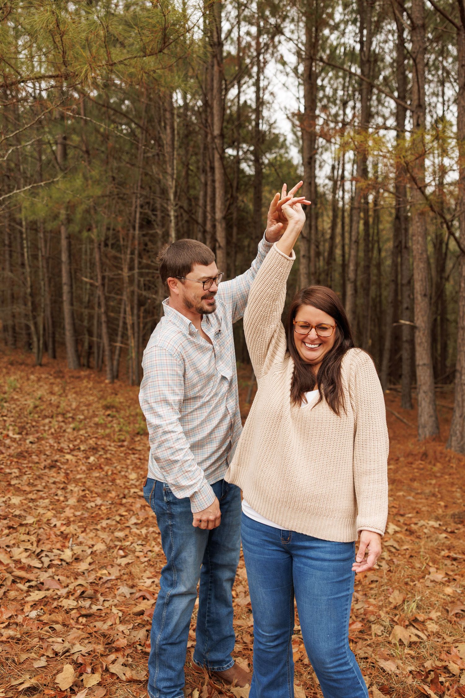 A couple shares playful moments together while standing among fallen autumn leaves in a pine forest.