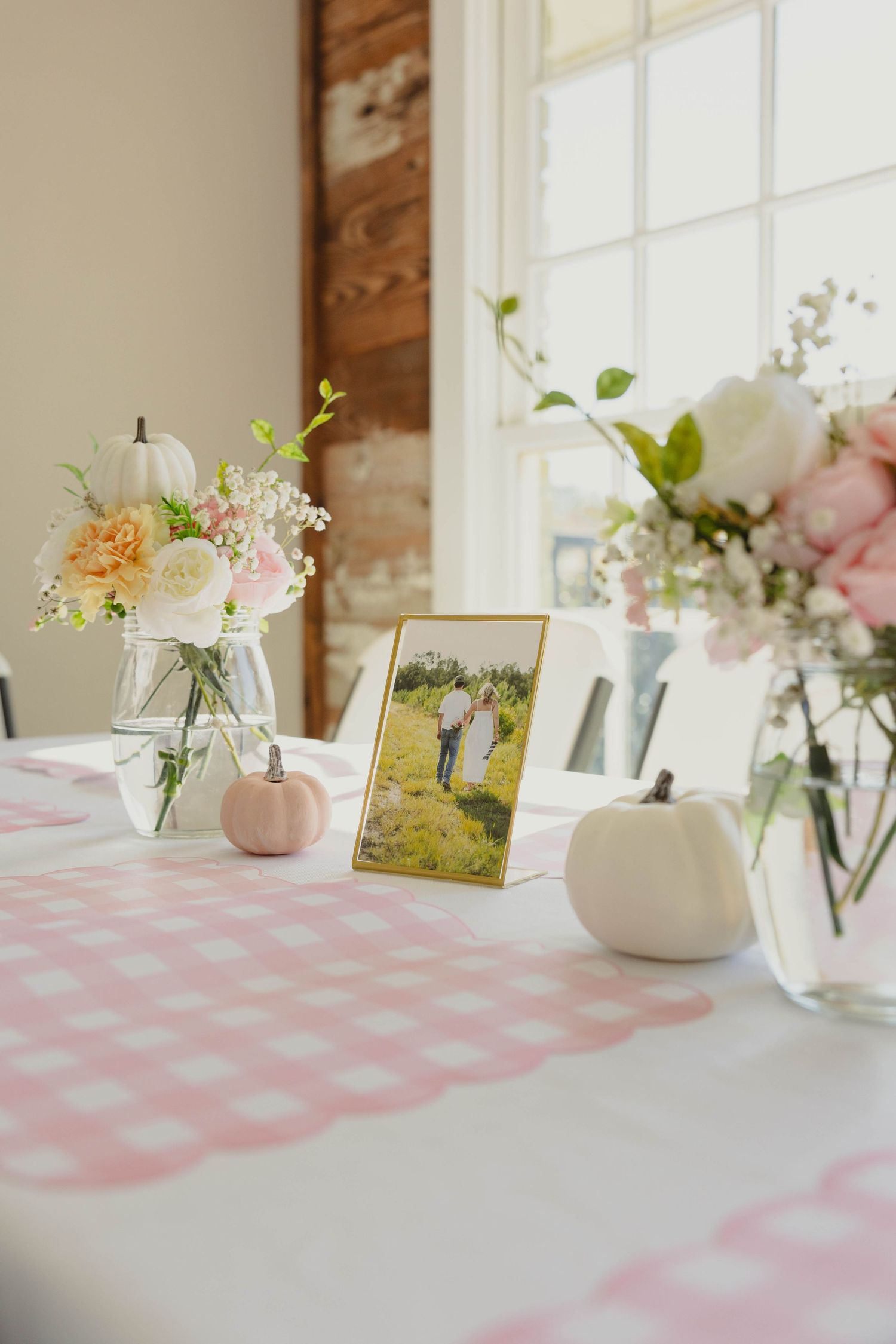 A pink gingham tablecloth with delicate floral arrangements in white vases and a framed photo on a sunlit table.
