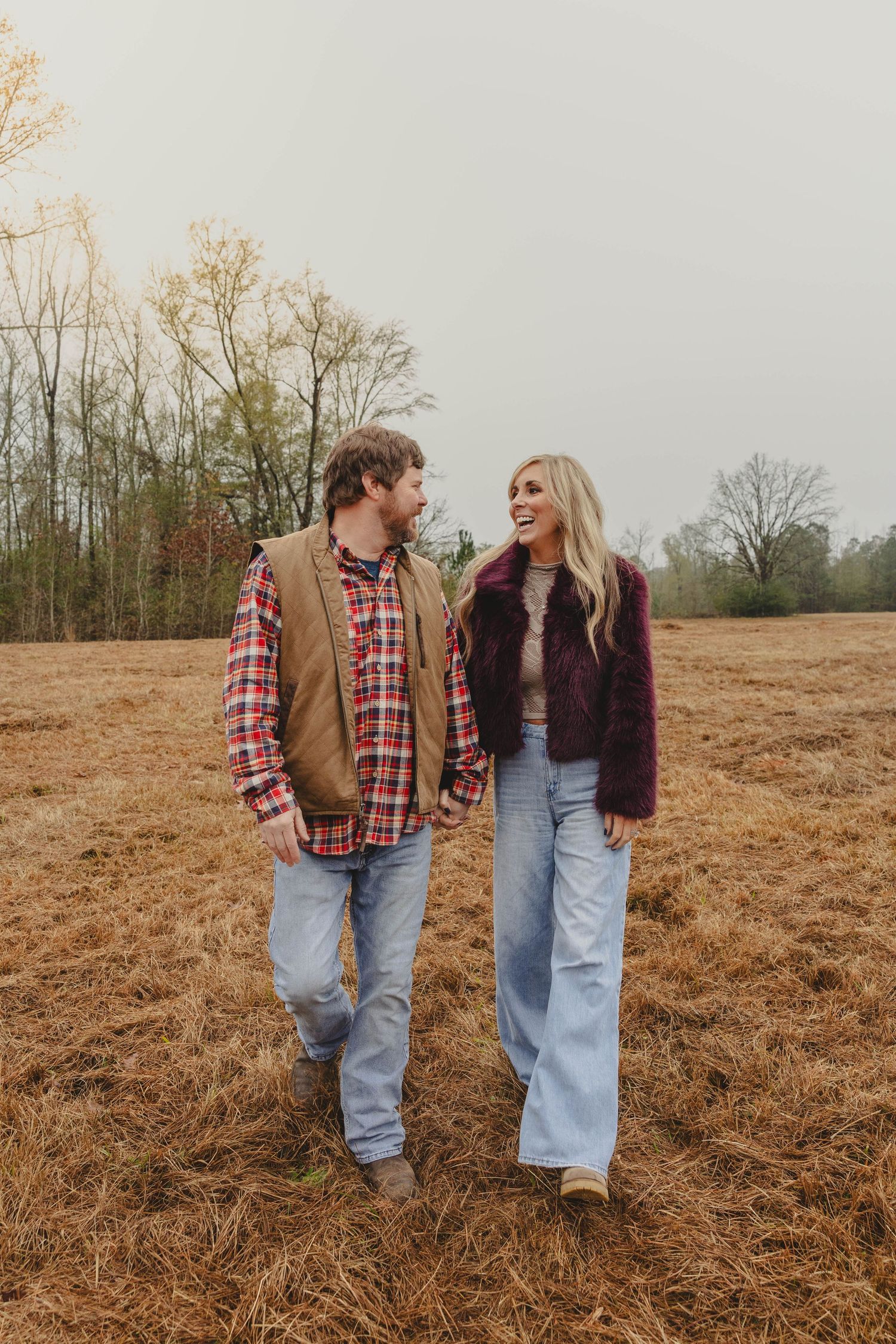 A couple in matching plaid shirts and jeans walk together through a golden autumn field on an overcast day.