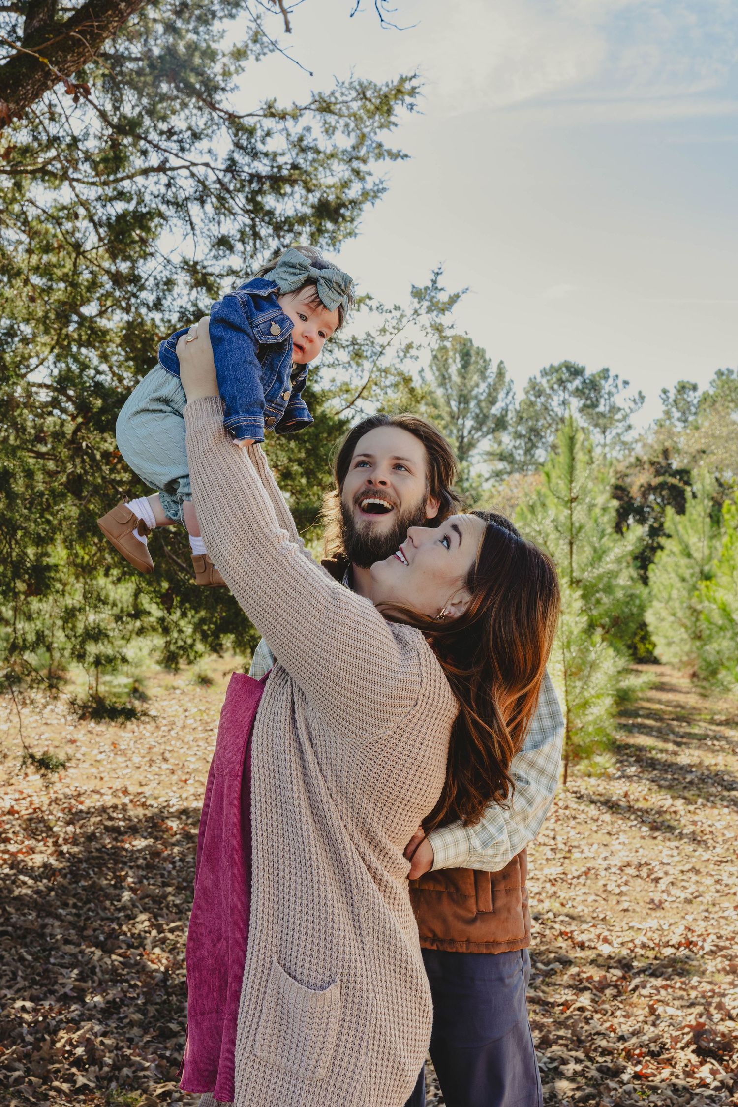 A person wearing a beige sweater lifts a small child playfully in an autumn outdoor setting with trees in the background.
