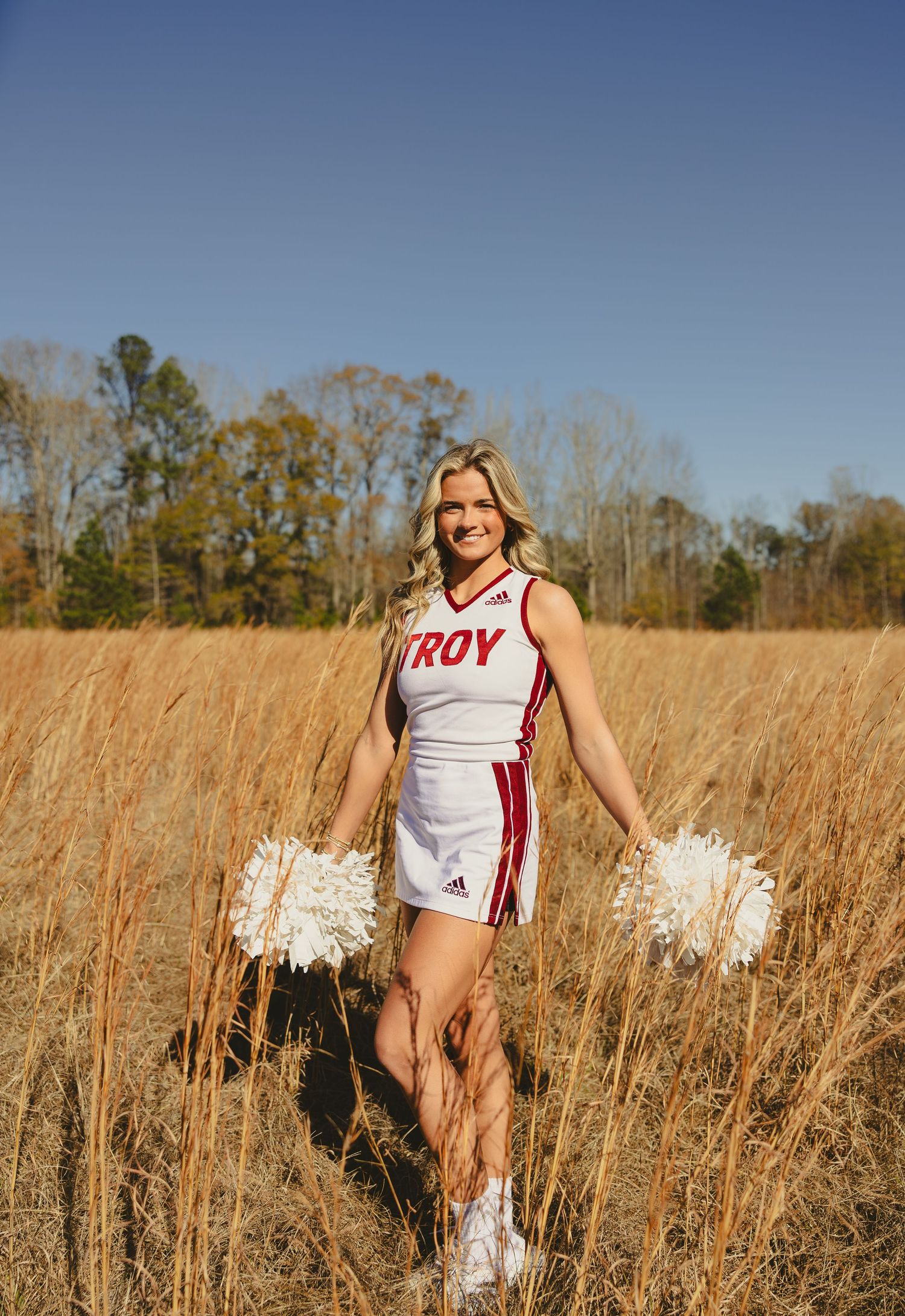 A cheerleader in a Troy uniform holds white pom poms while standing in a golden autumn field on a sunny day.