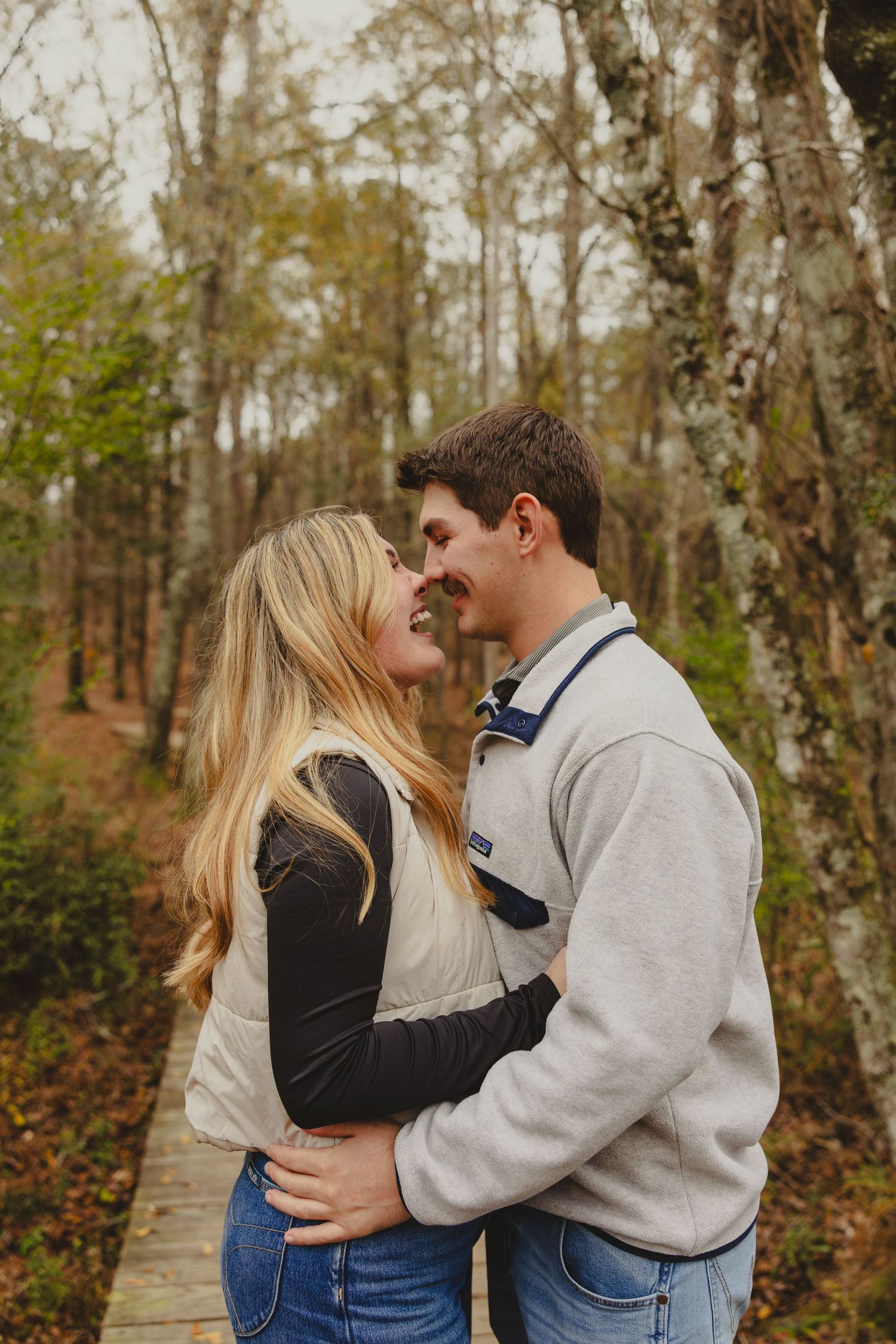 A couple shares intimate romantic moments in a wooded autumn setting while walking along a forest path.