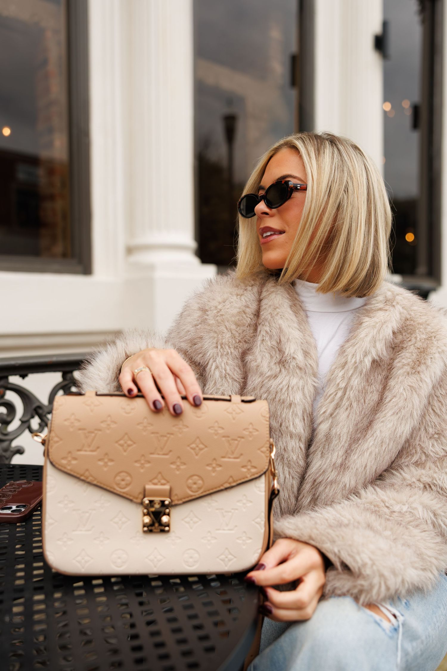 A woman in a fluffy beige fur coat holds a Louis Vuitton monogram embossed handbag while sitting outdoors.