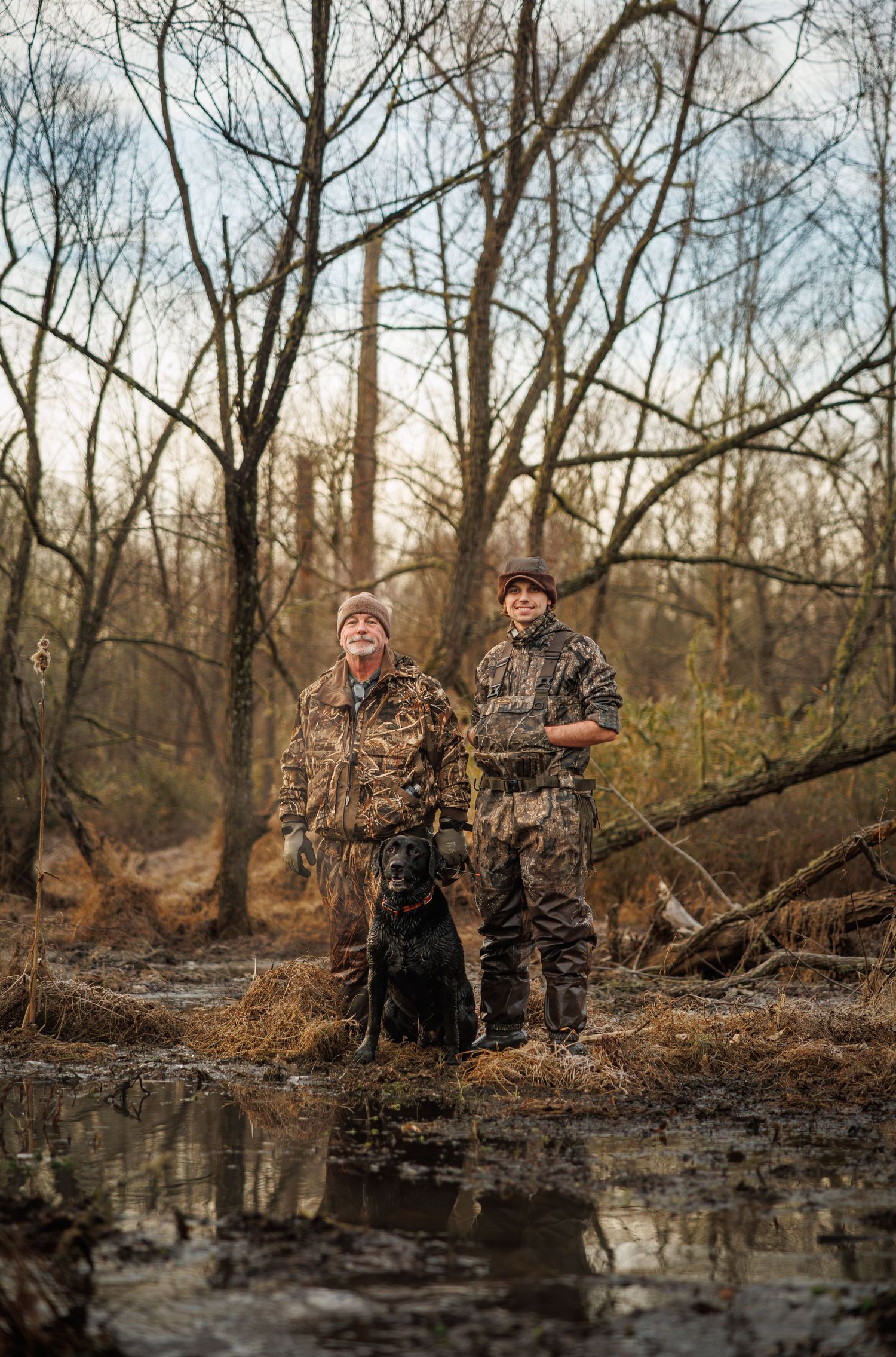 Two hunters in camouflage gear stand with their black dog by a reflective pond in a winter forest setting.