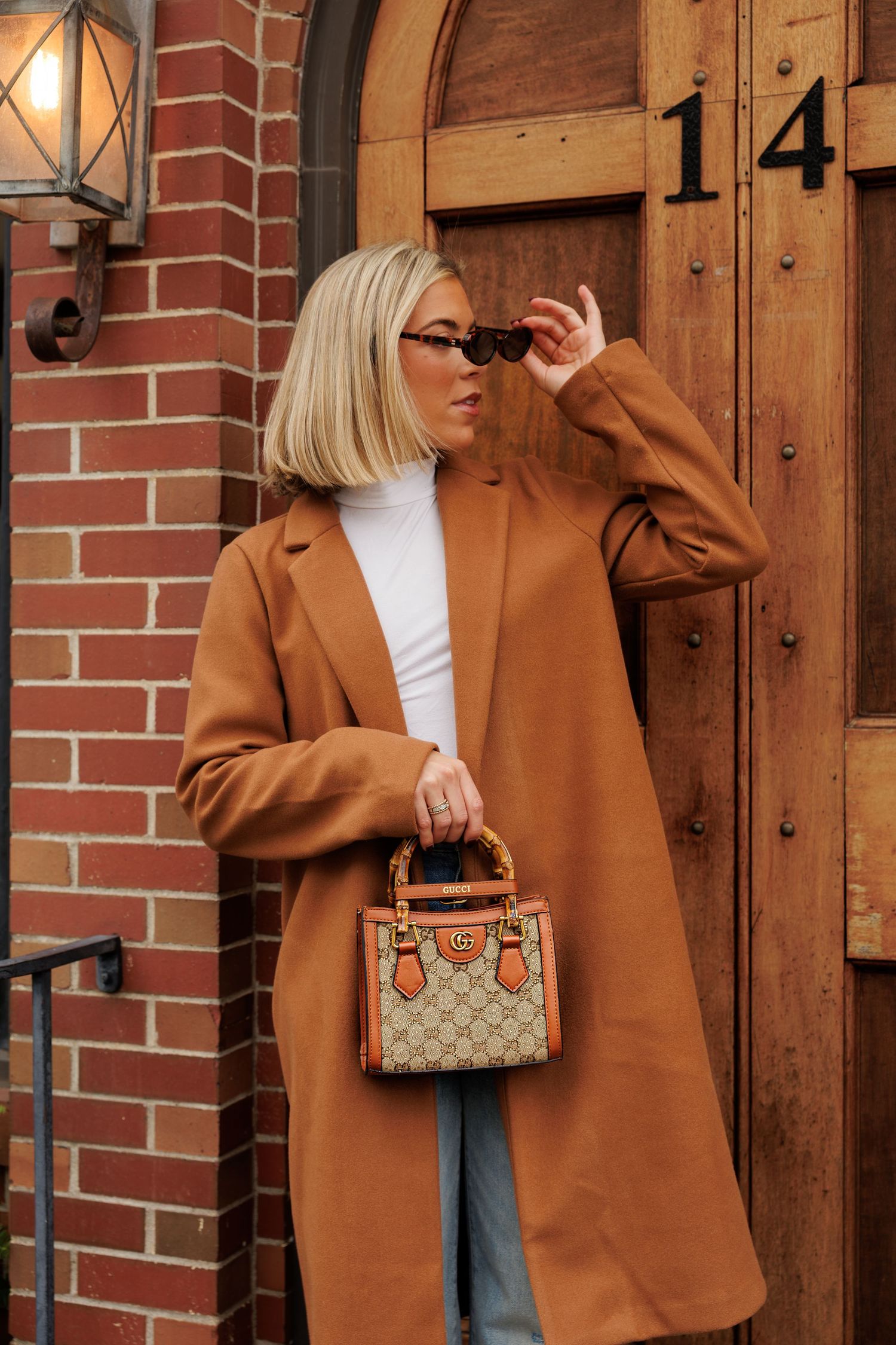 Woman in camel coat and jeans poses against brick wall with designer handbag in multiple stylish shots.