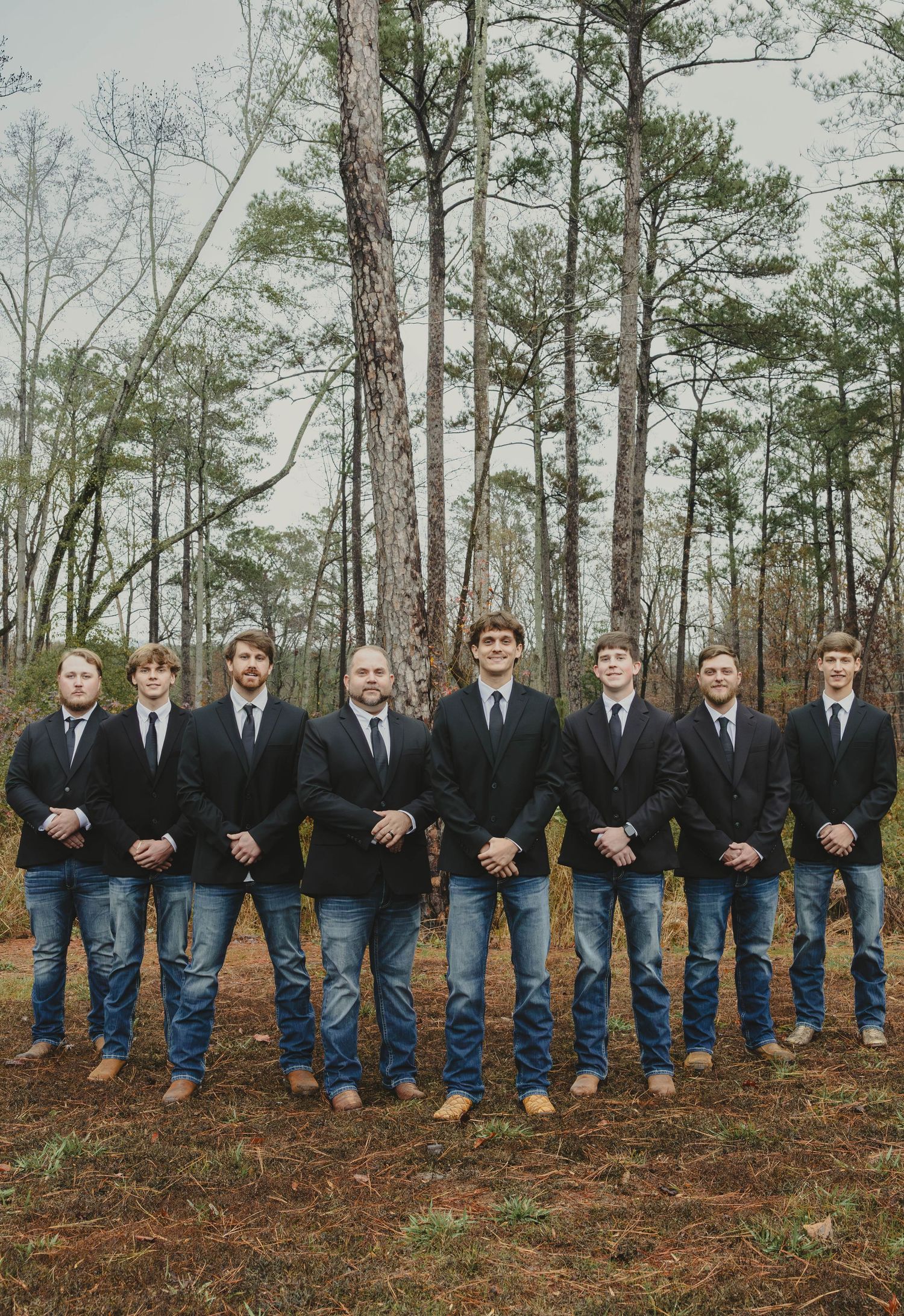 Groomsmen in black suits and blue jeans sit in a row on pine needles in a wooded outdoor setting.