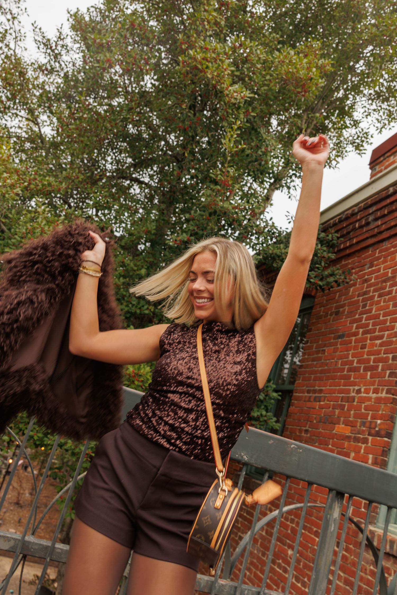 Person in a brown dress raises their arms joyfully while posing outdoors against a brick wall and trees.