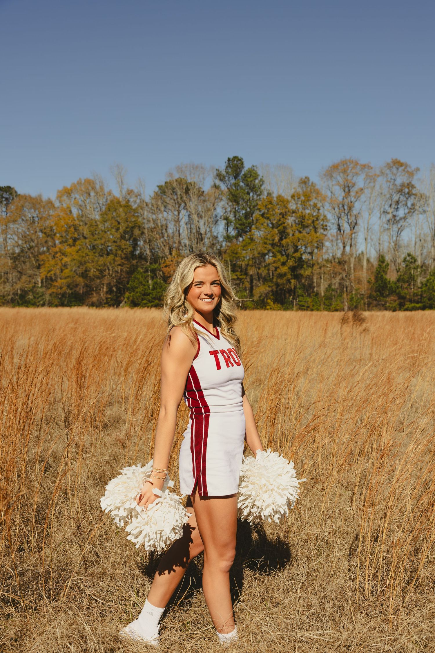 Cheerleader poses in a field of tall golden grass while holding white pom poms.
