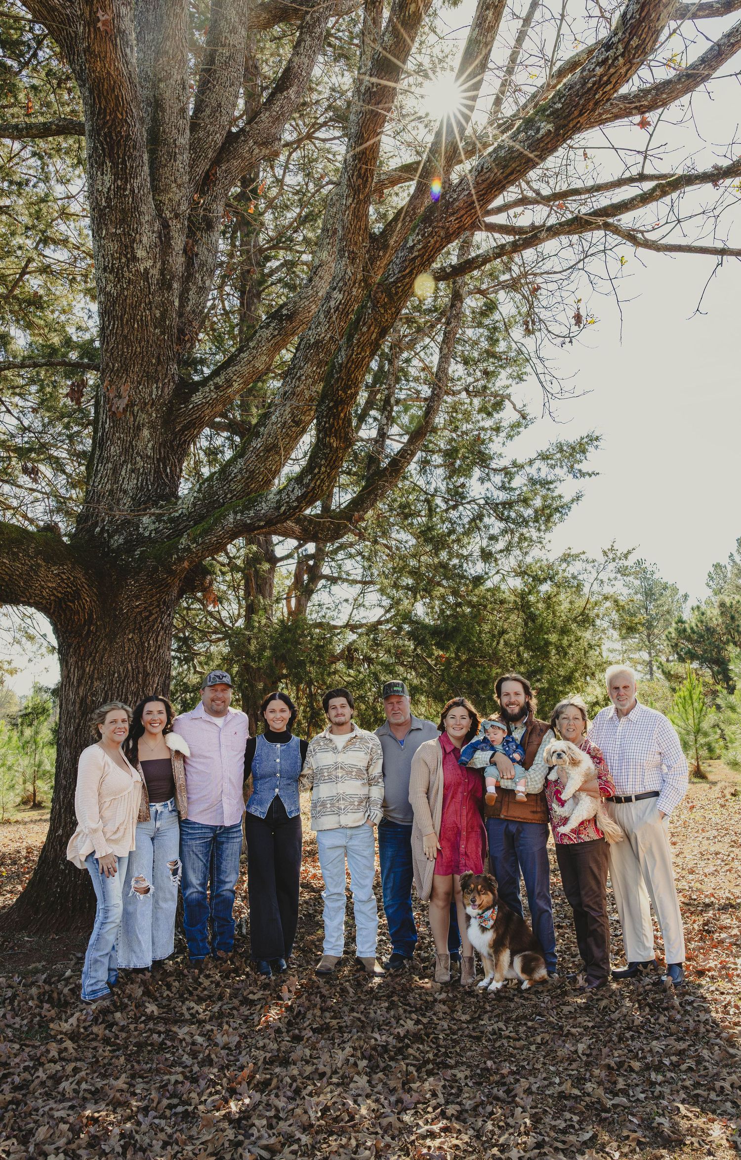 A group of people gather for an outdoor family photo session under large trees in natural lighting.