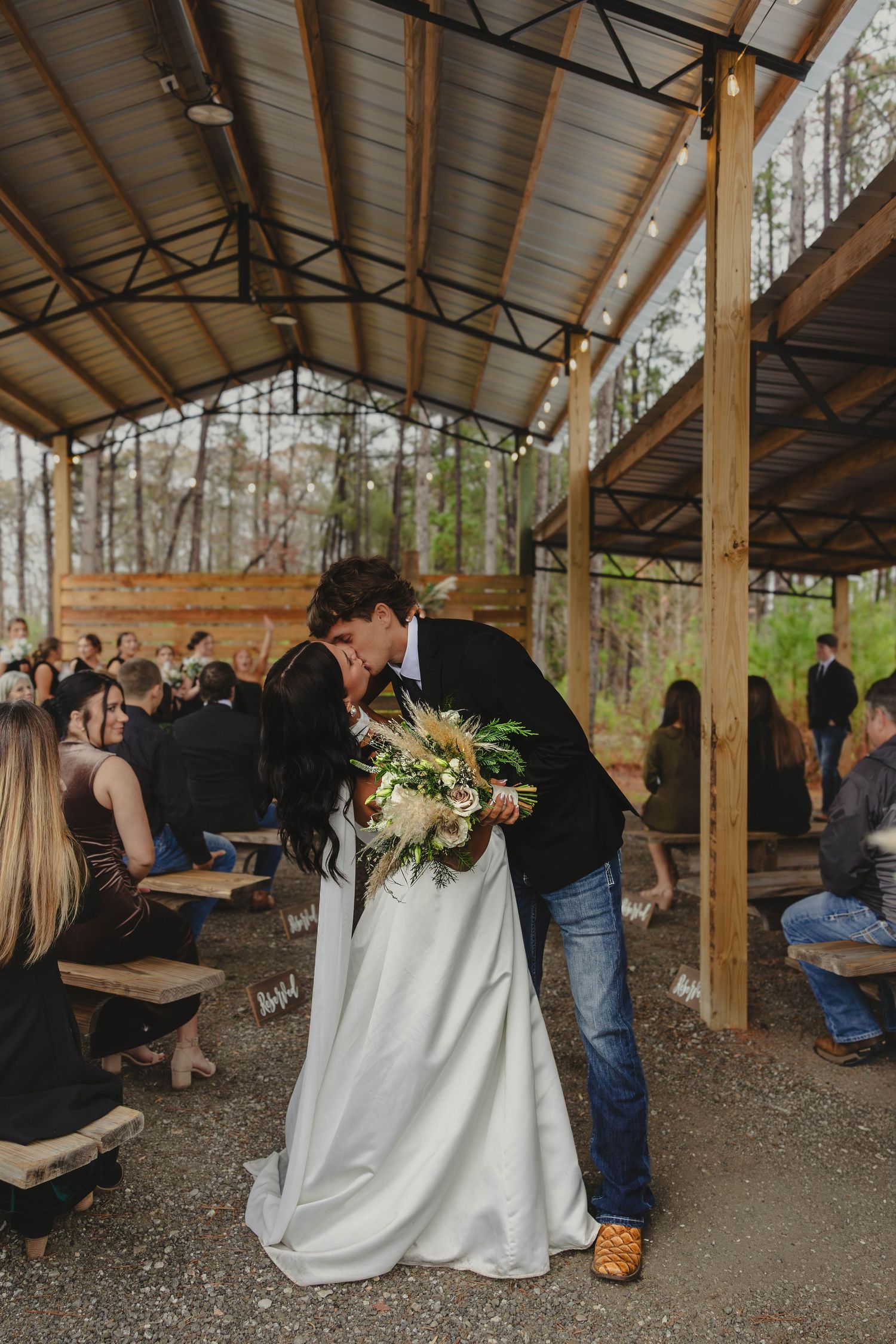 Wedding reception scene in a rustic wooden pavilion with guests mingling under exposed beams and natural lighting.