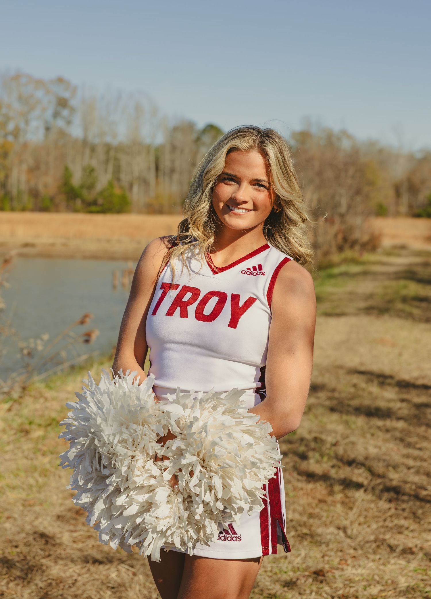 A cheerleader in a white and red Troy uniform poses with pom poms outdoors by a lake on a sunny day.