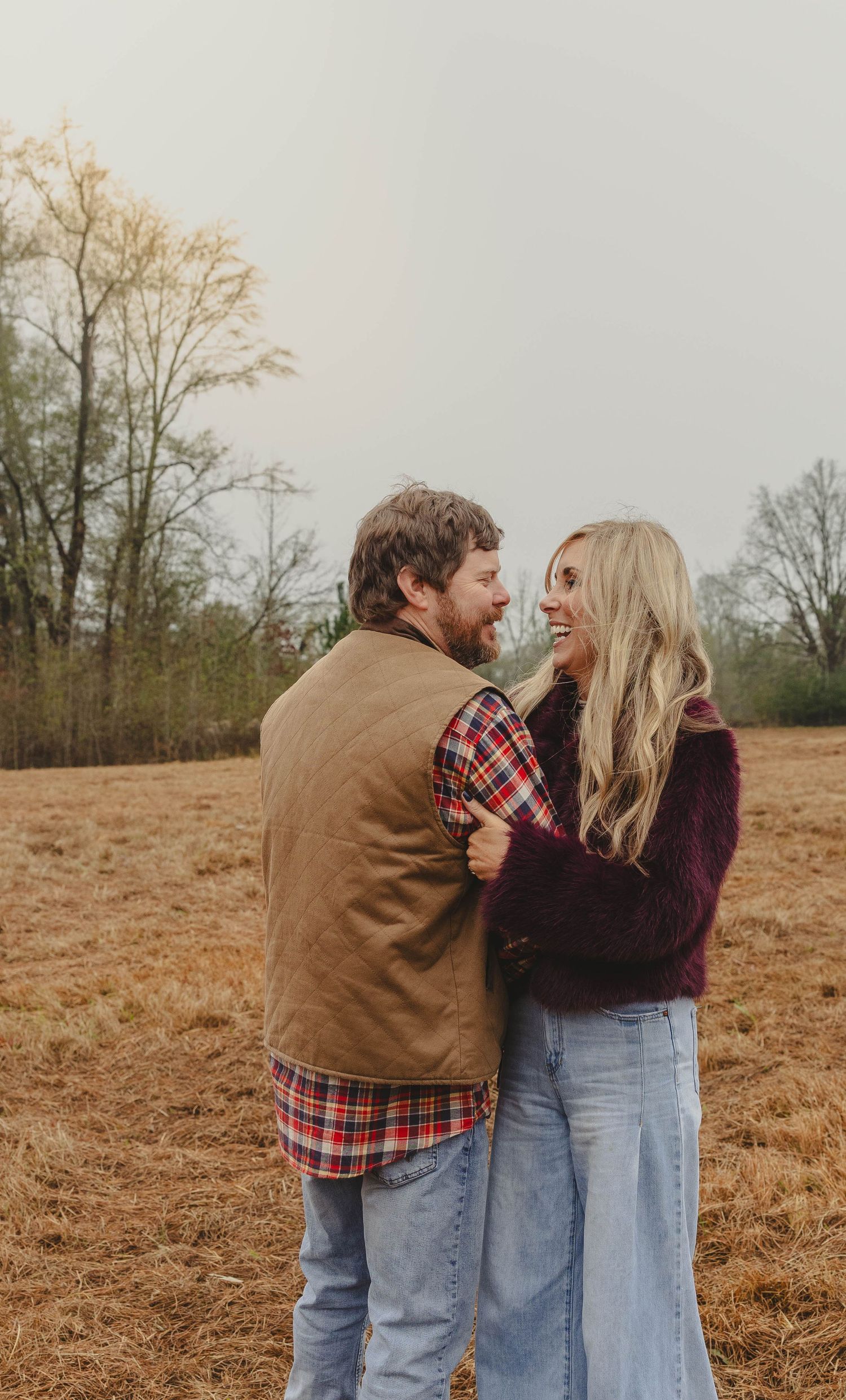 A couple shares a playful moment together in a rustic autumn field wearing casual denim and plaid attire.
