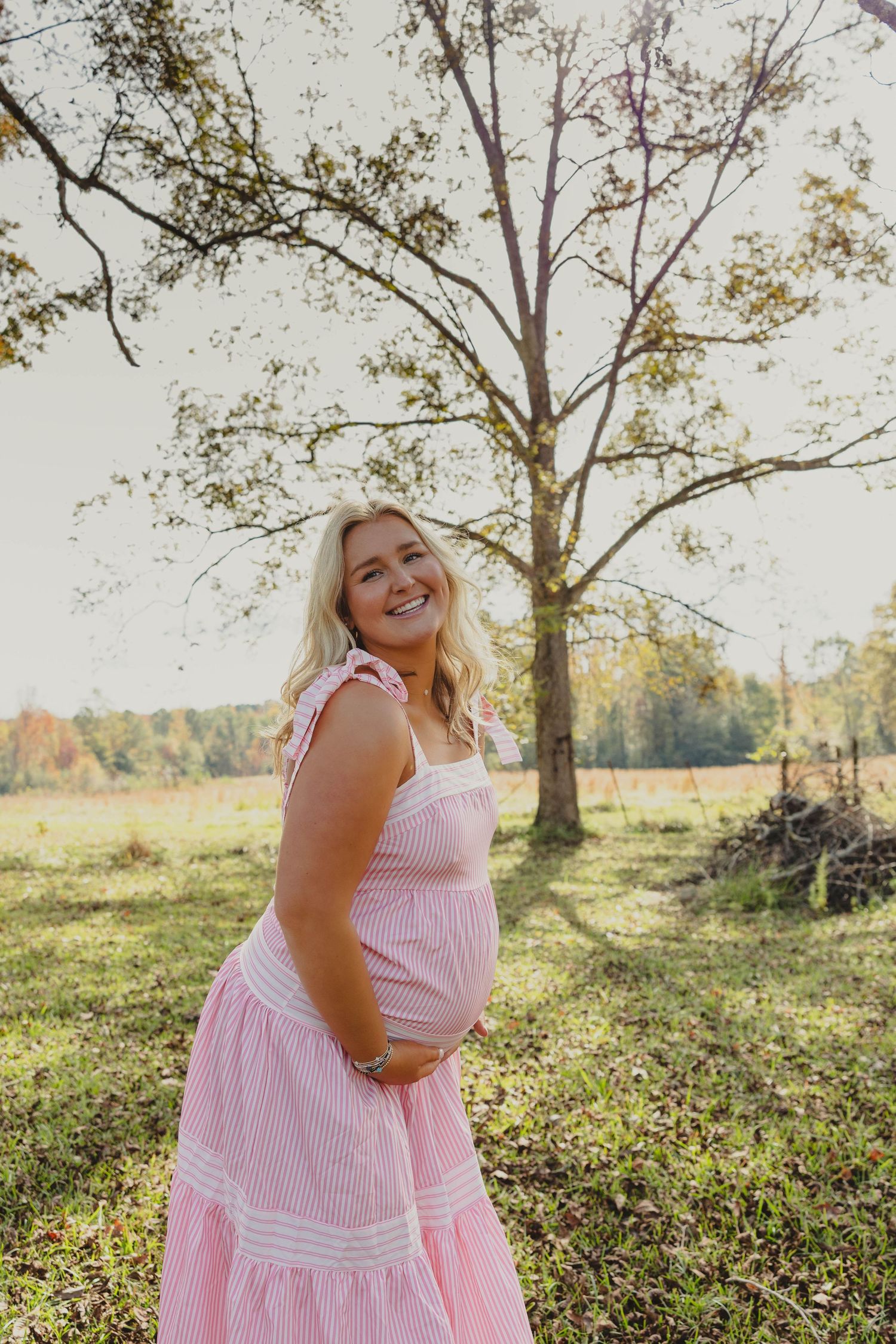A person in a pink and white striped dress poses joyfully in a sunny meadow with trees in the background.