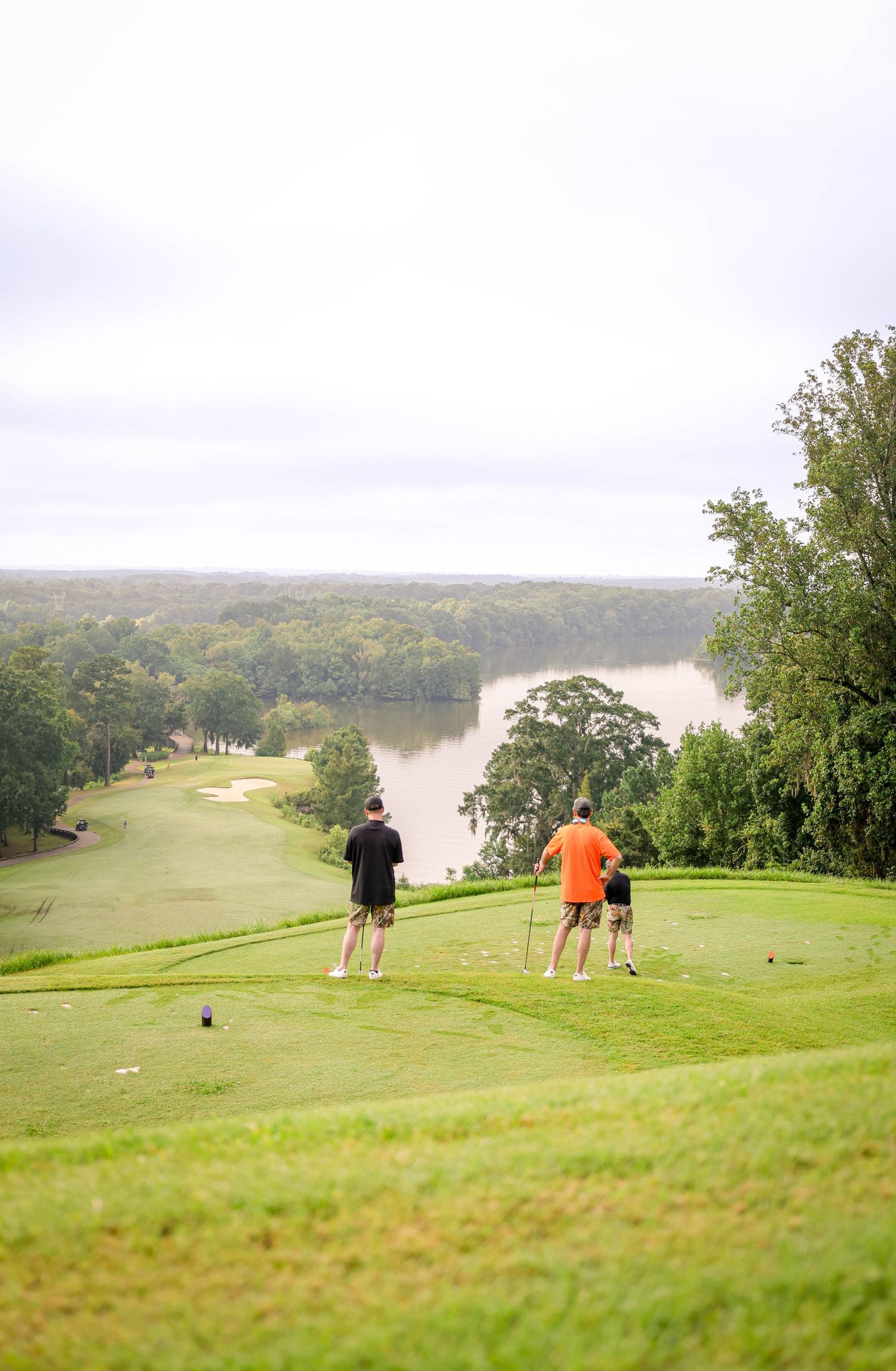 Golfers play on a scenic hillside course overlooking a lake on a sunny day.