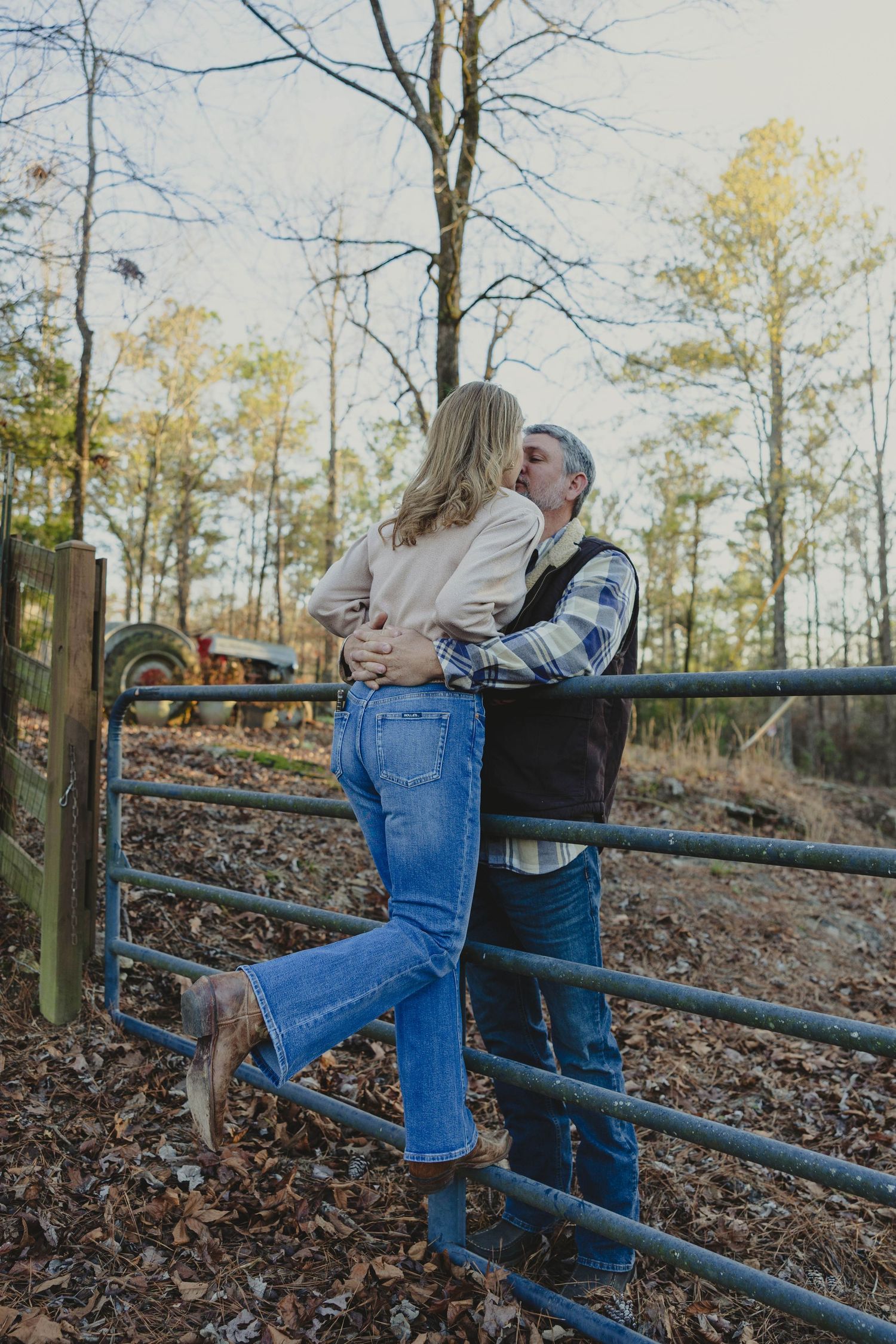 A couple in casual attire leans playfully against a rustic fence surrounded by autumn leaves in a wooded area.