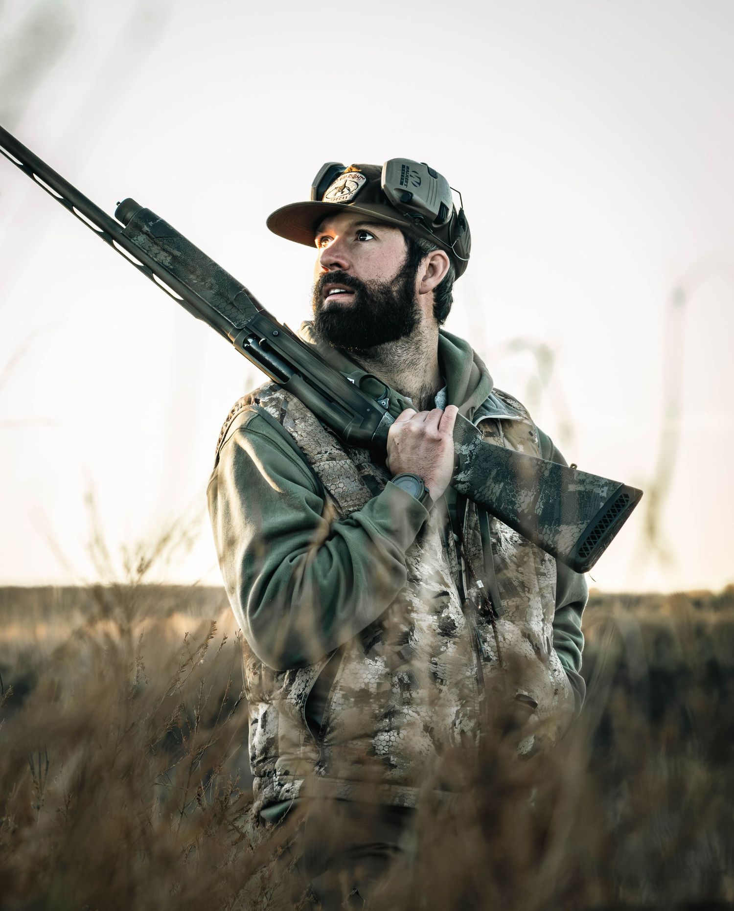 A hunter in camouflage gear aims a rifle while crouching in tall grass during golden hour.