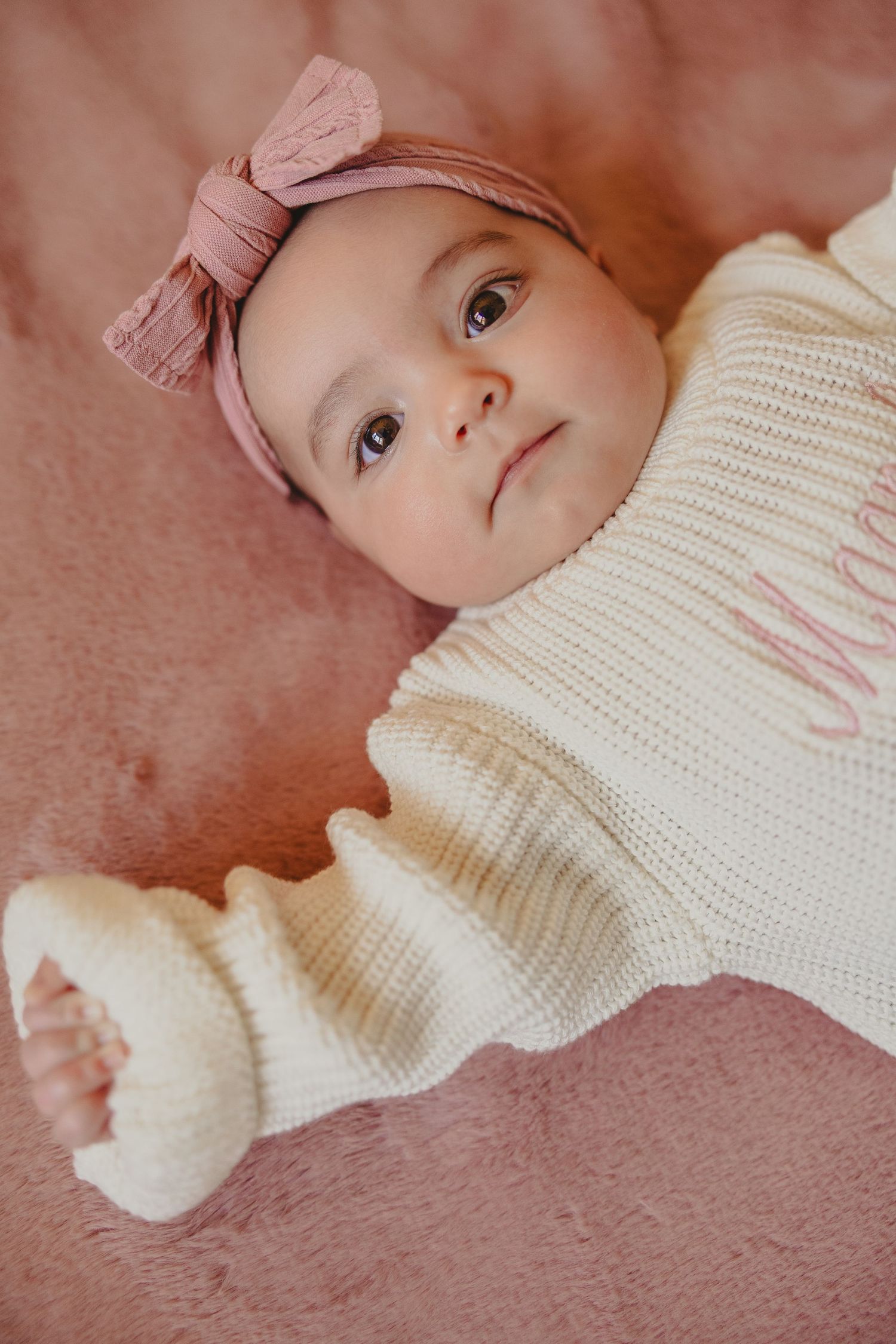 Baby wearing cream knit sweater lies on pink textured surface looking upward with curious expression.