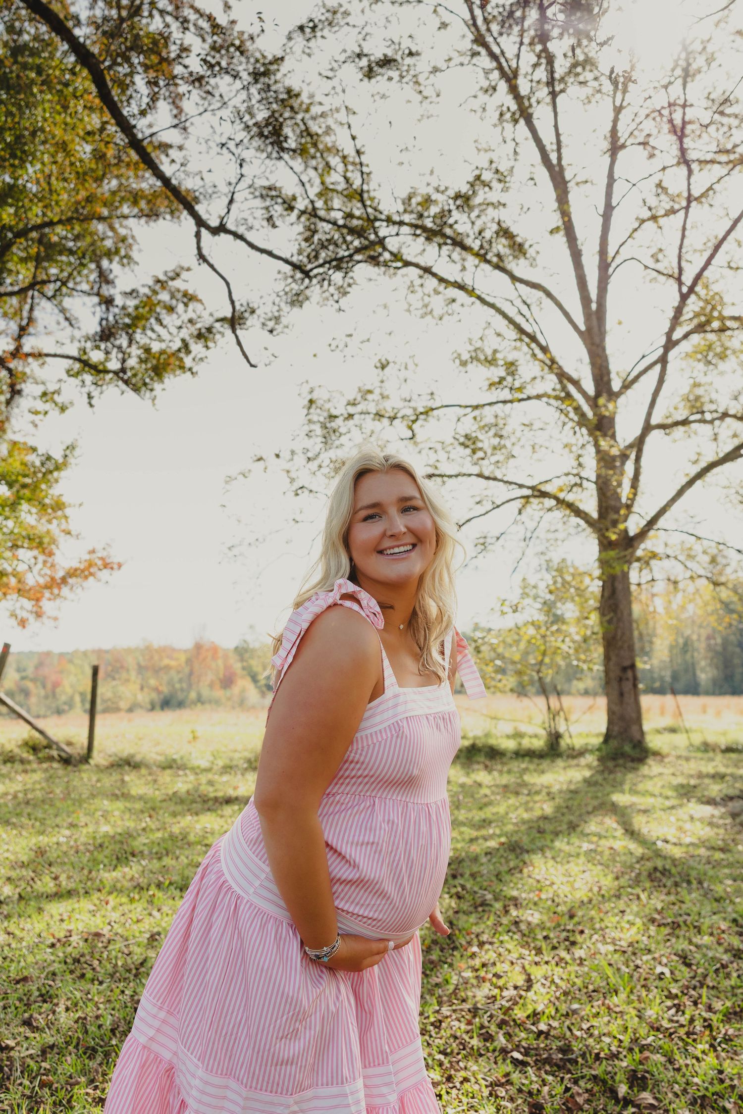 Person in pink striped dress posing in a sunlit autumn field with trees in the background.