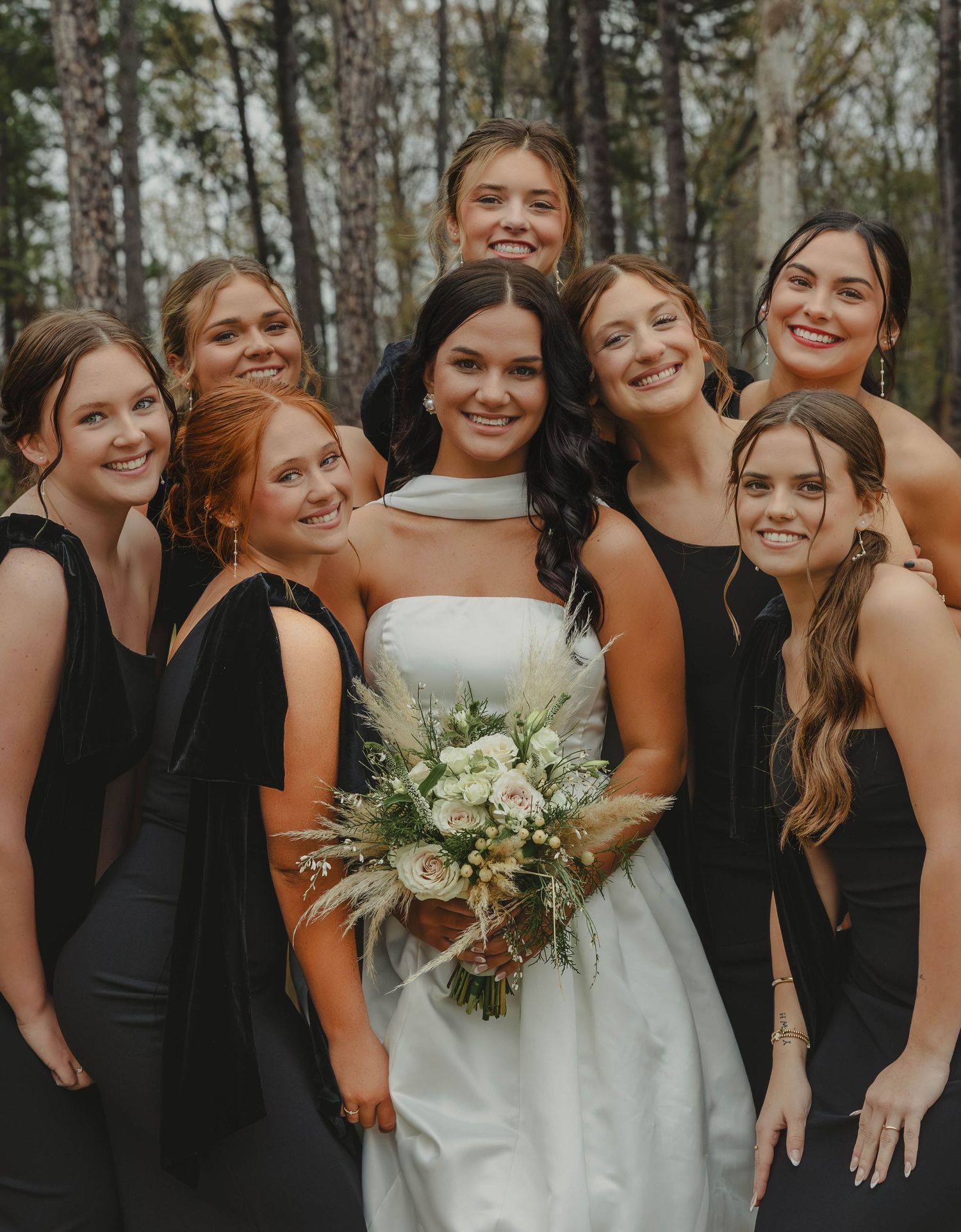 A bridal party in black dresses surrounds a bride in white holding a neutral-toned bouquet in a wooded setting.