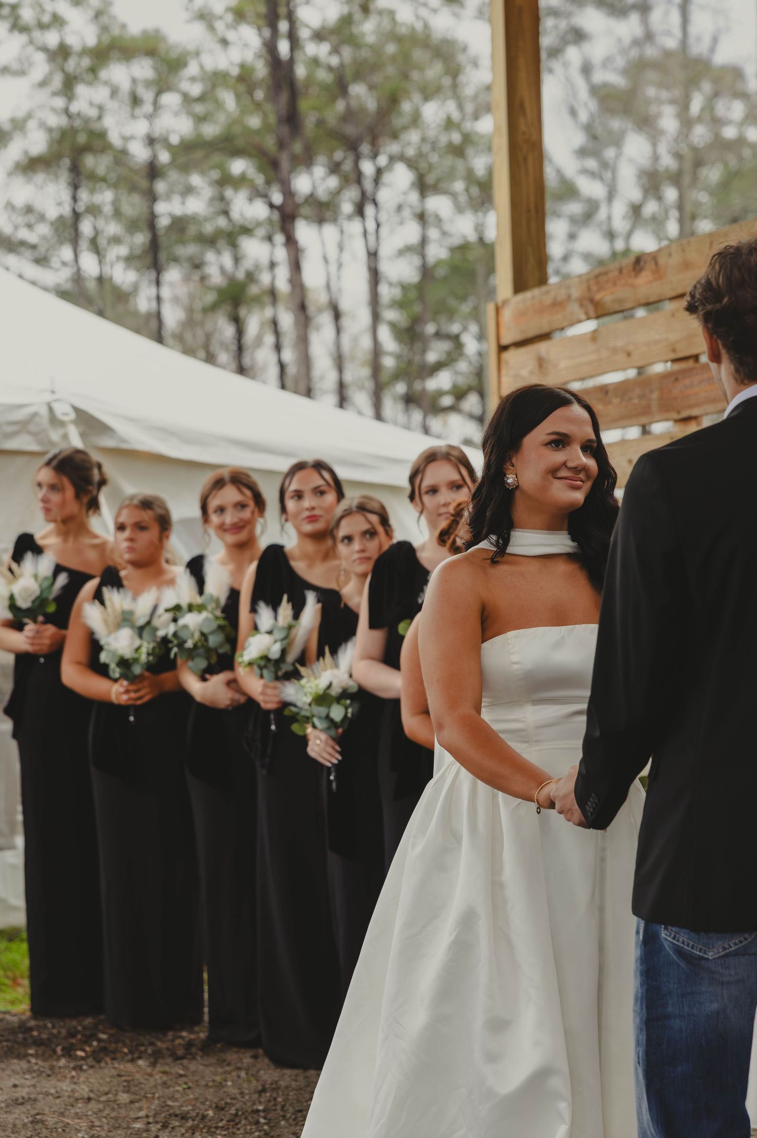 Wedding party in black bridesmaid dresses stands in a line outdoors with pine trees in the background.