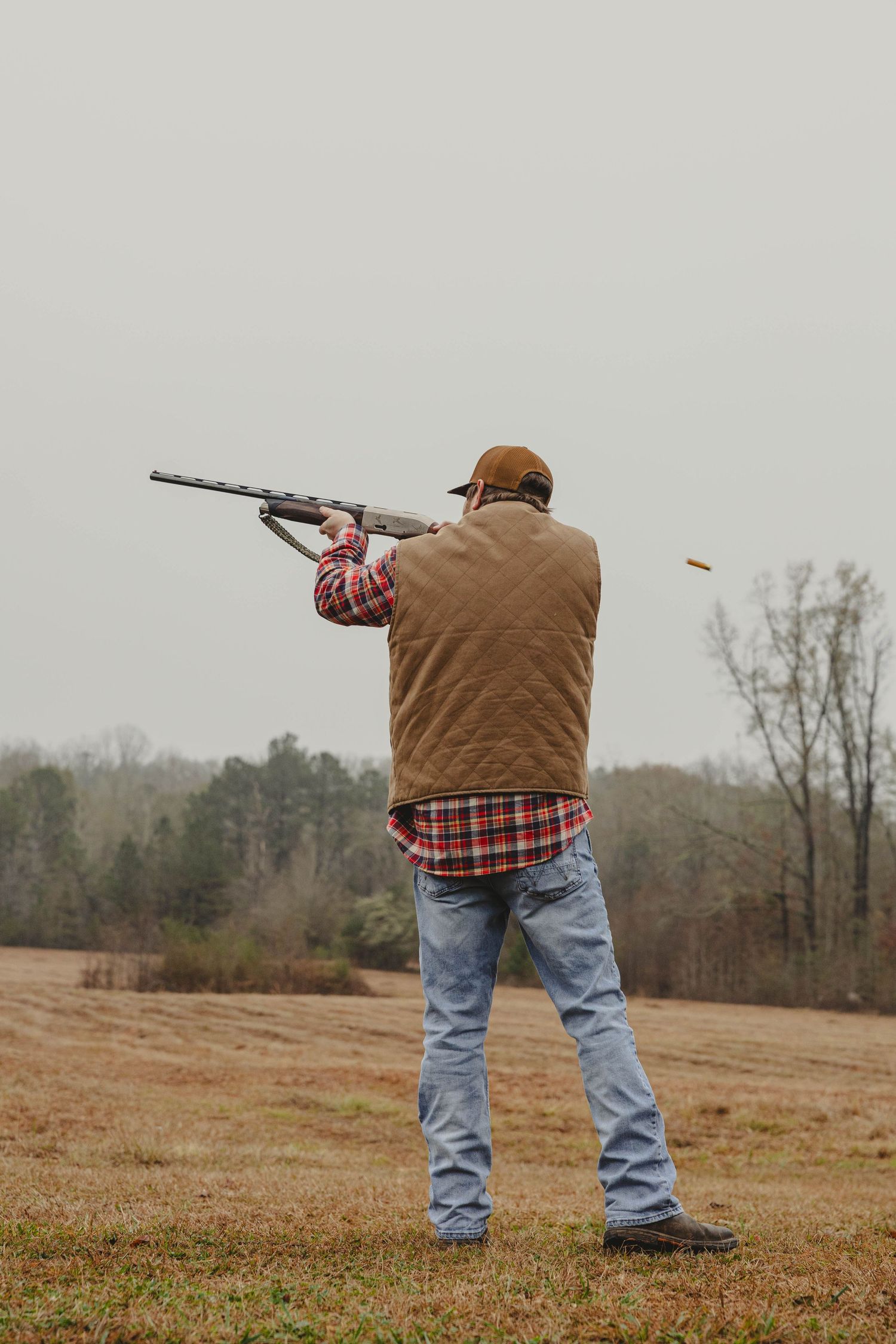 A person in jeans and brown jacket aims a shotgun across an open field during target practice.