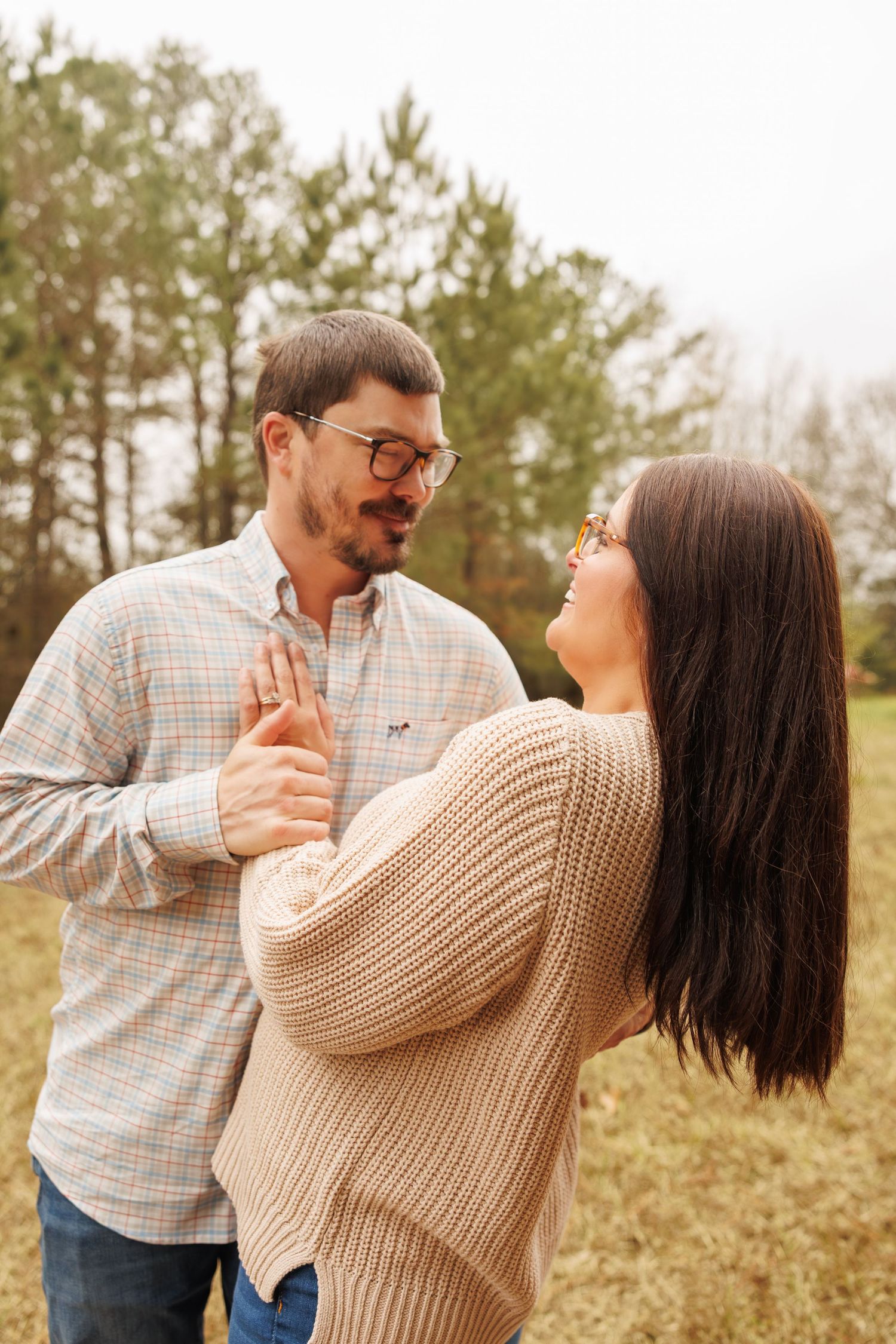 A couple in casual sweater and plaid shirt share a romantic moment outdoors against a backdrop of pine trees.