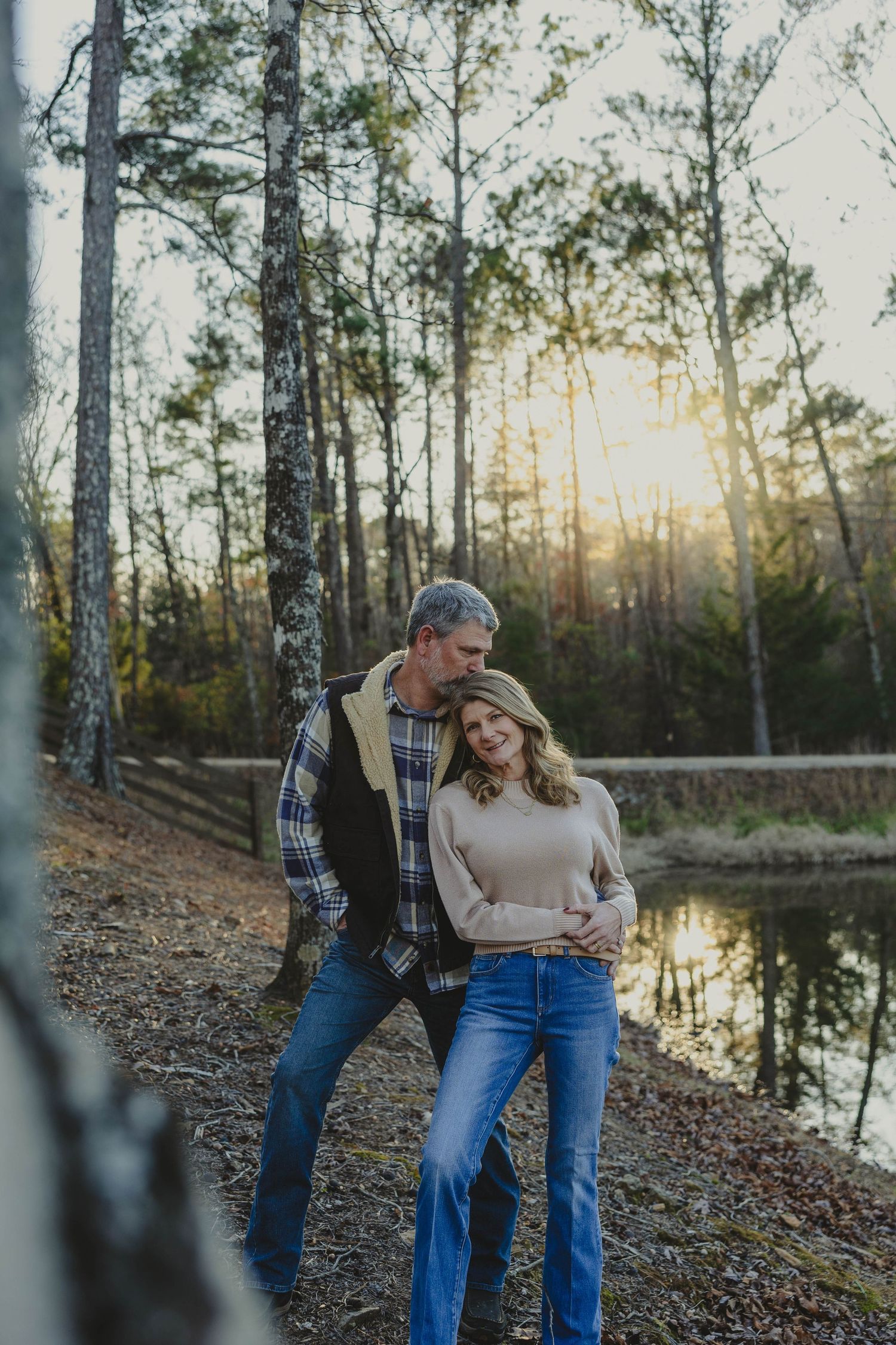 A couple shares a playful moment by a serene lake at sunset, surrounded by tall trees.
