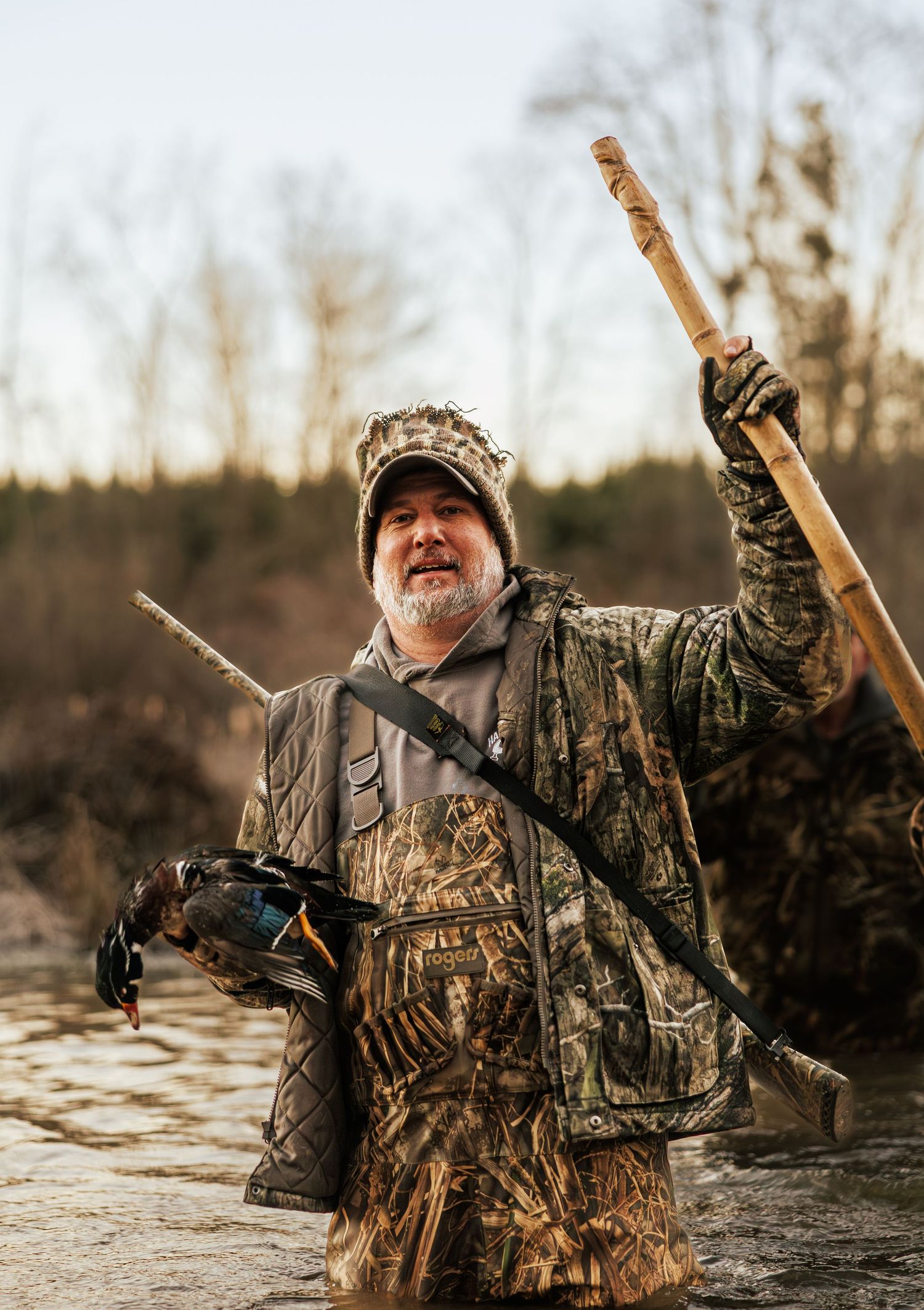 A hunter in camouflage gear stands in water holding hunting equipment during a winter expedition.