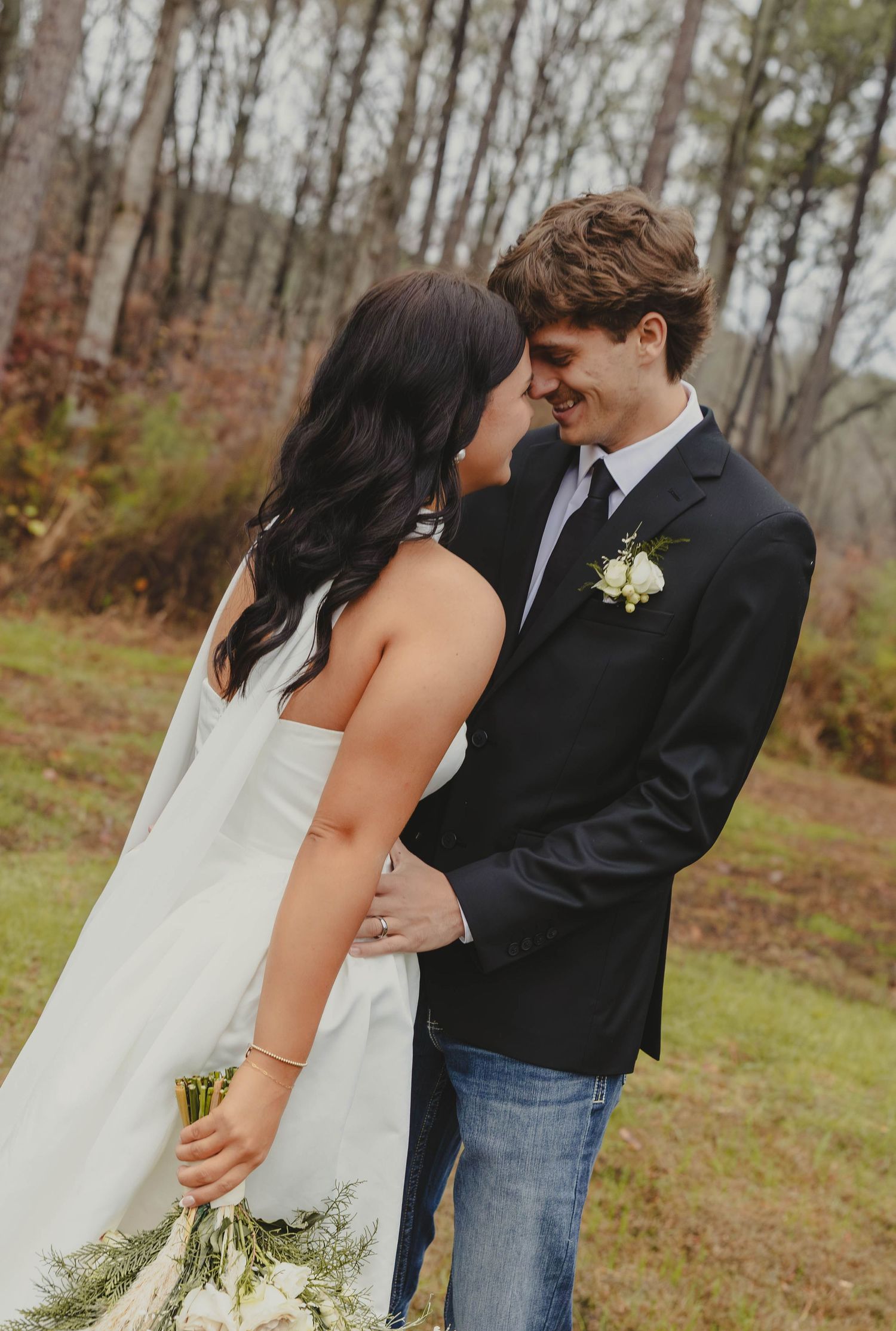 A couple shares an intimate moment in their wedding attire outdoors with autumn trees in the background.