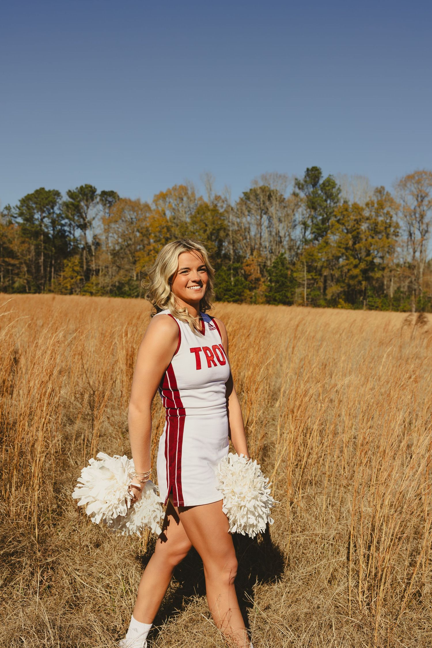 Cheerleader in white uniform with pom poms performs routine in autumn field with golden grass and trees in background.
