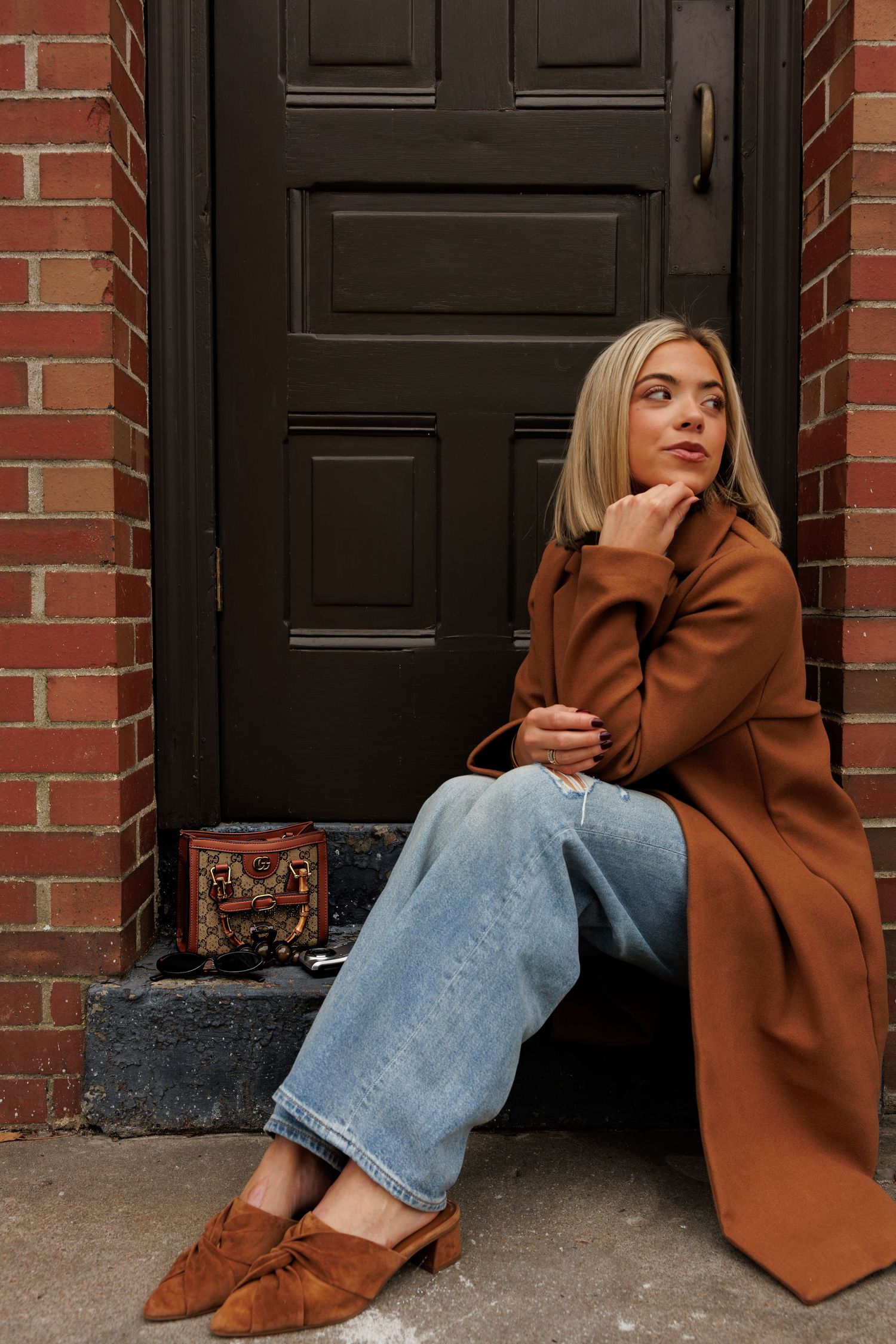 Stylish autumn look with camel coat, blue jeans and brown suede shoes against brick building entrance.