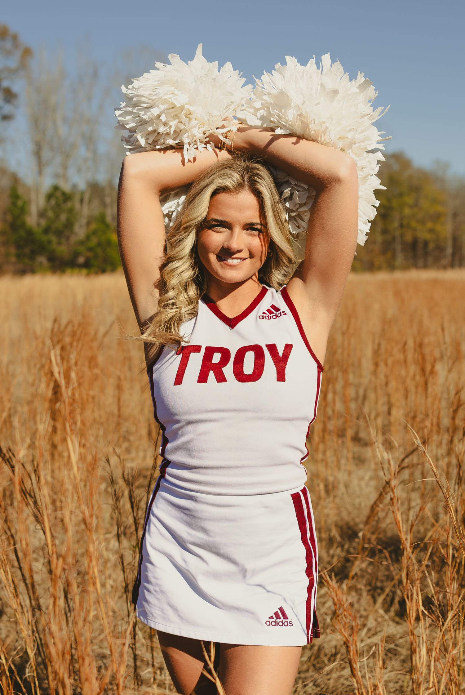 Cheerleader in white Troy University uniform poses in tall grass field during autumn photoshoot.