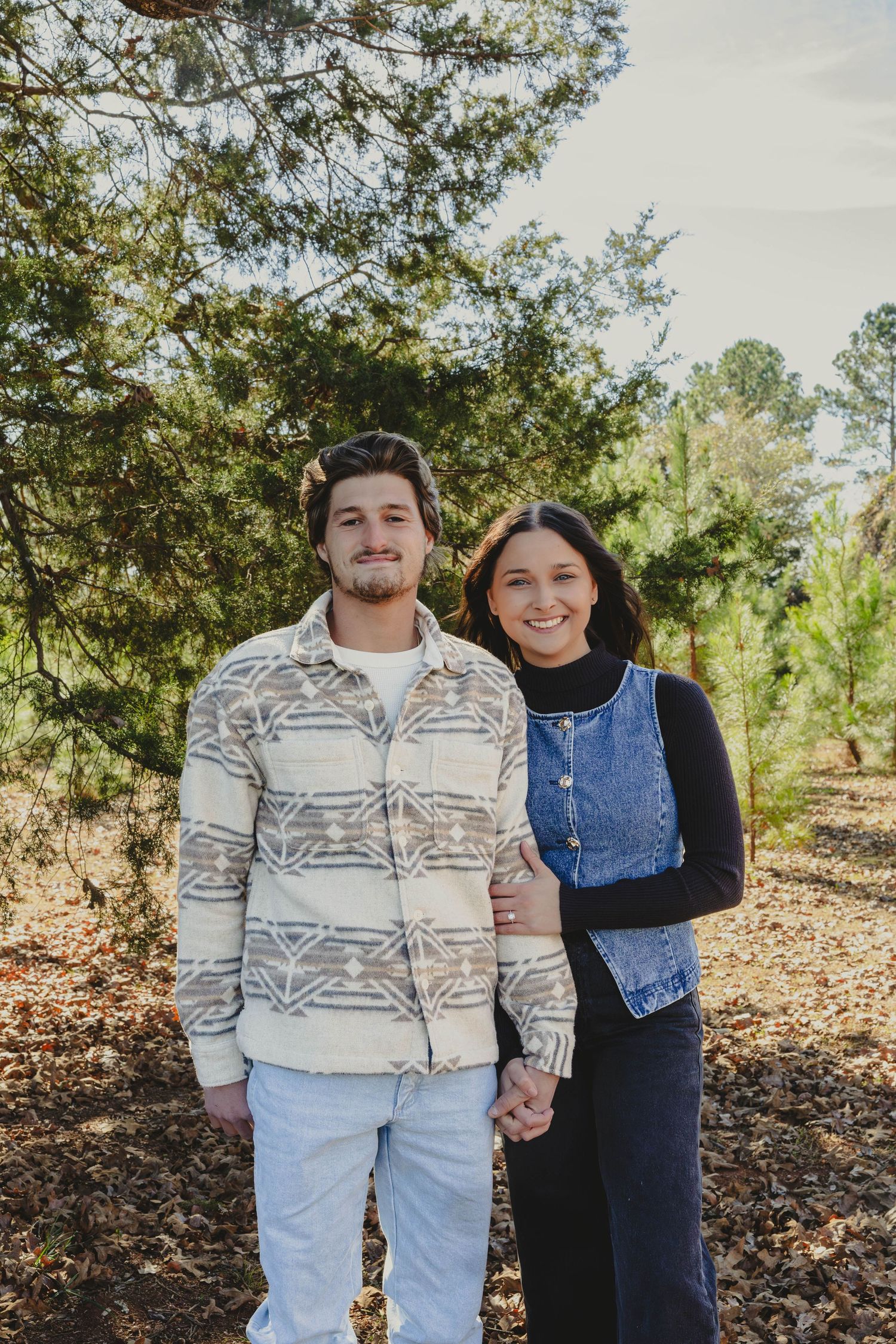 A couple in coordinating outfits stands together outdoors among autumn leaves and trees.