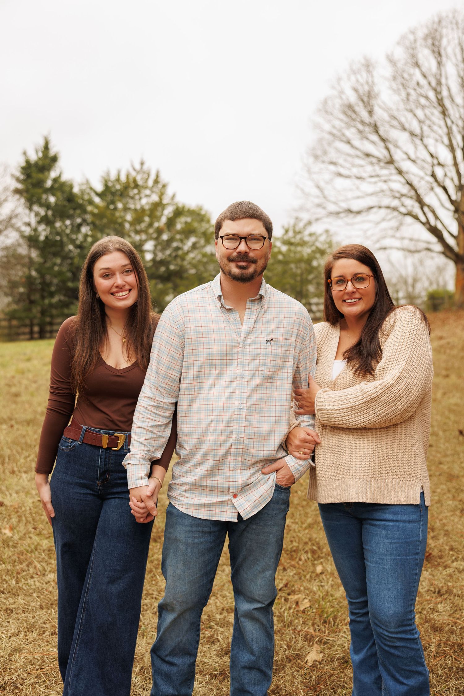Three people in casual fall attire standing together outdoors in a grassy field on an autumn day.