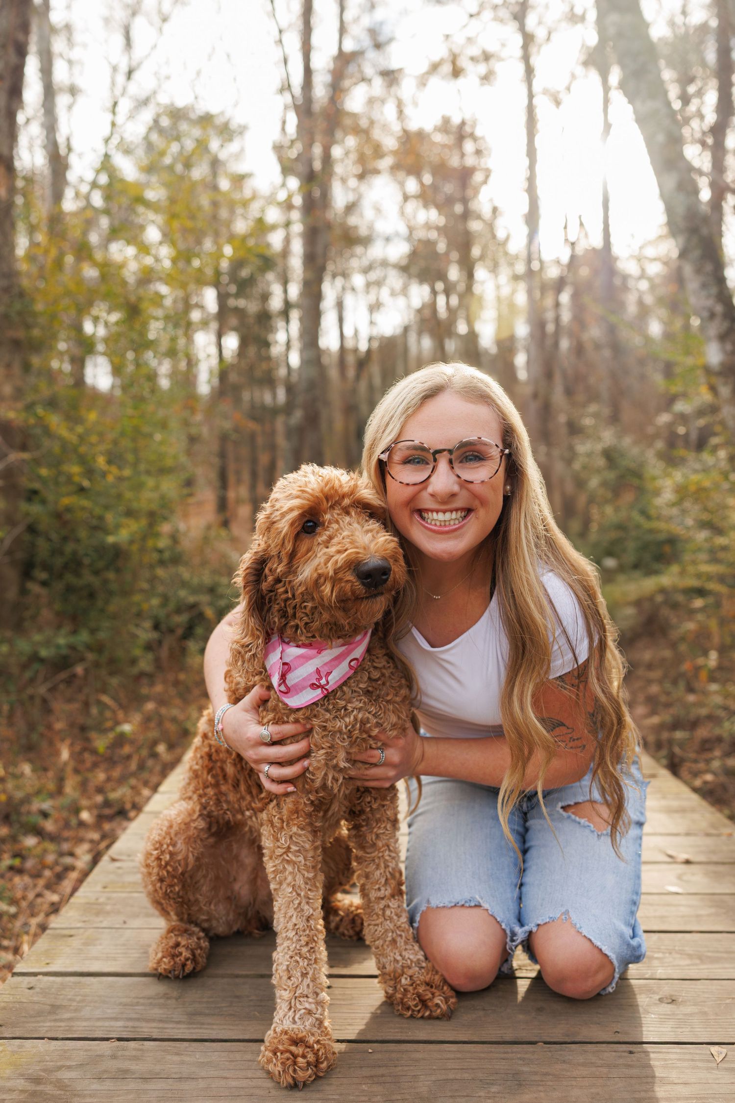 A happy goldendoodle dog with reddish-brown curly fur sits with someone on a wooden boardwalk in an autumn forest.