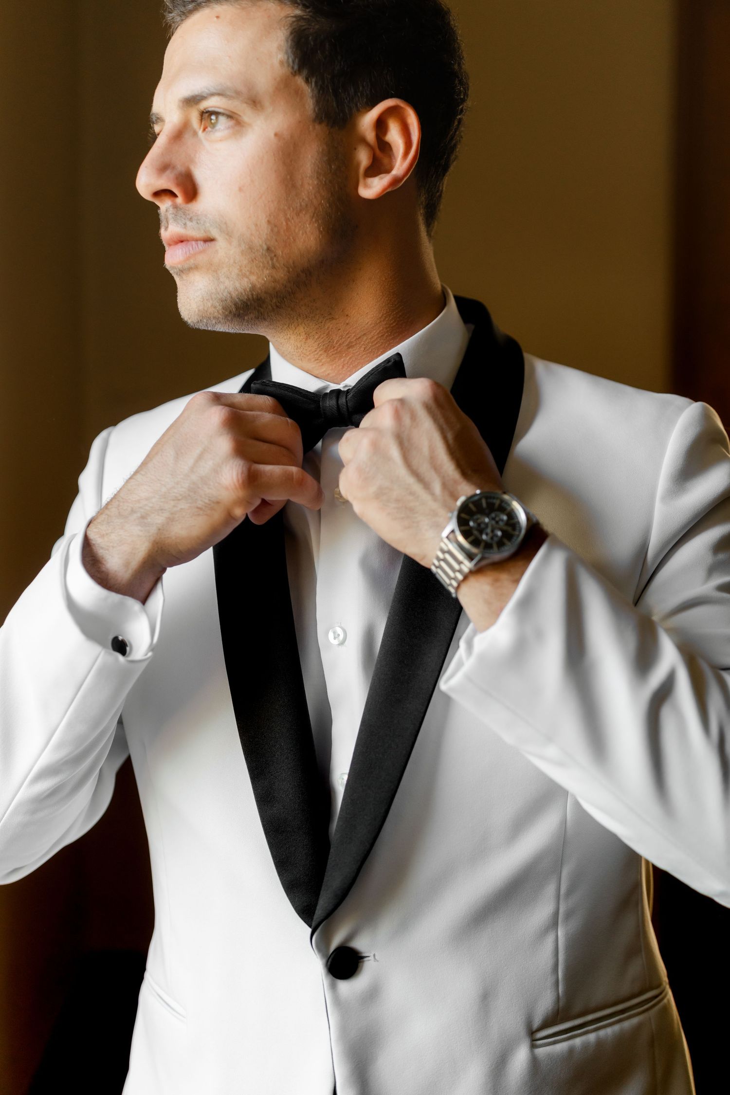 Groom in white tuxedo jacket and black bow tie adjusts his watch.