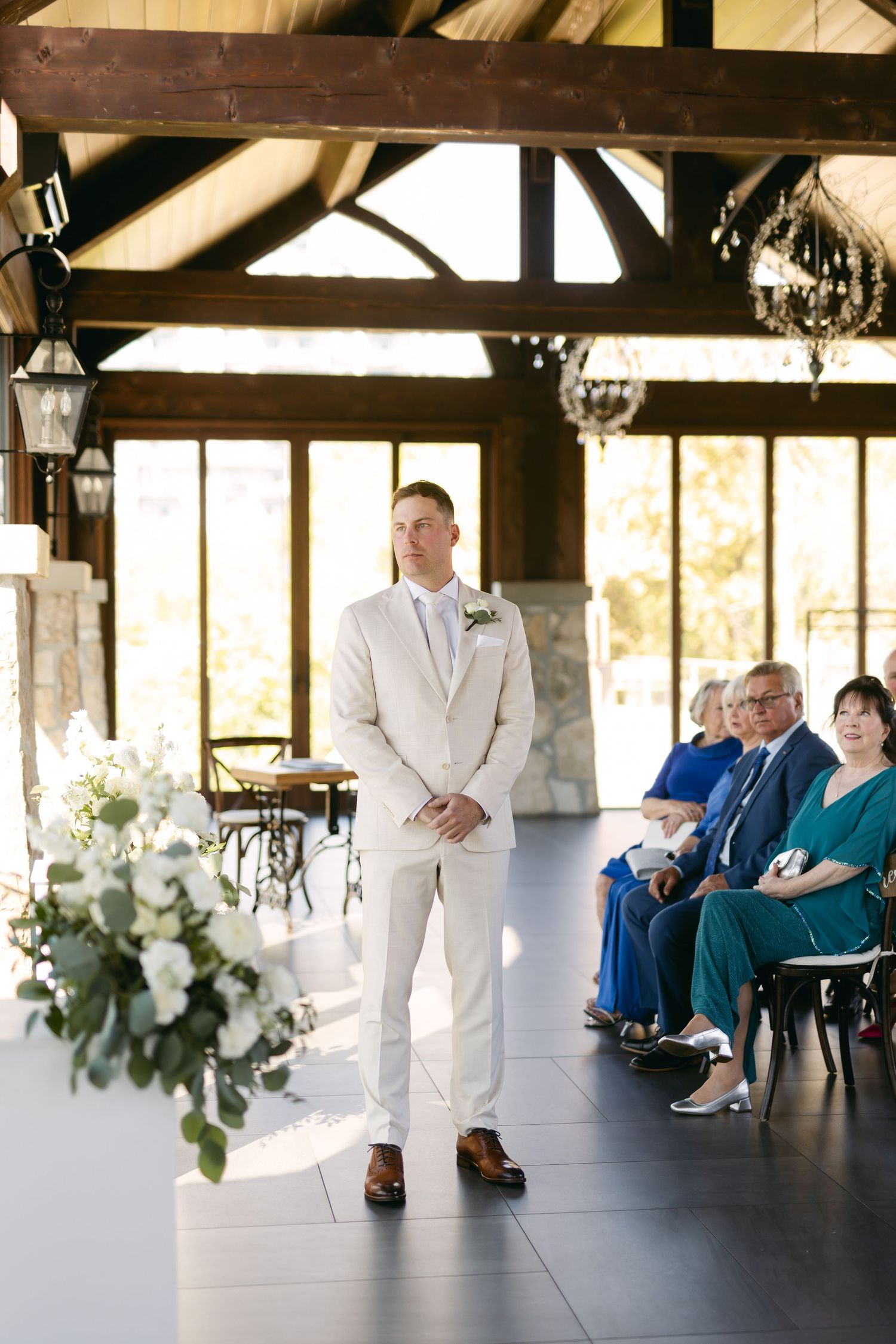 A groom in a light colored suit stands at the altar in a rustic venue with exposed wooden beams.