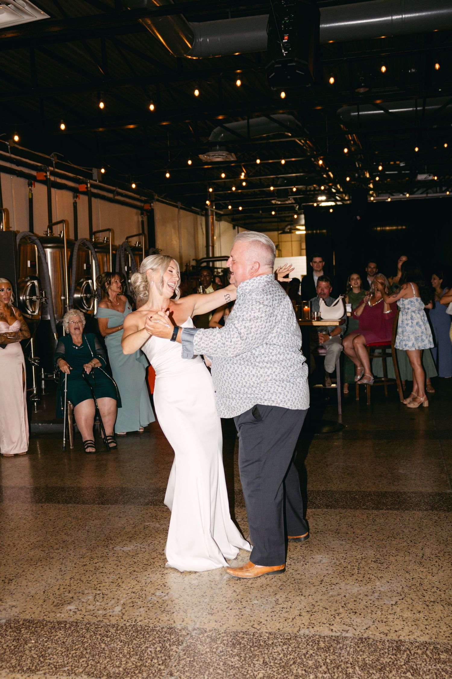 Wedding guests dancing together on polished floor under string lights at indoor reception.